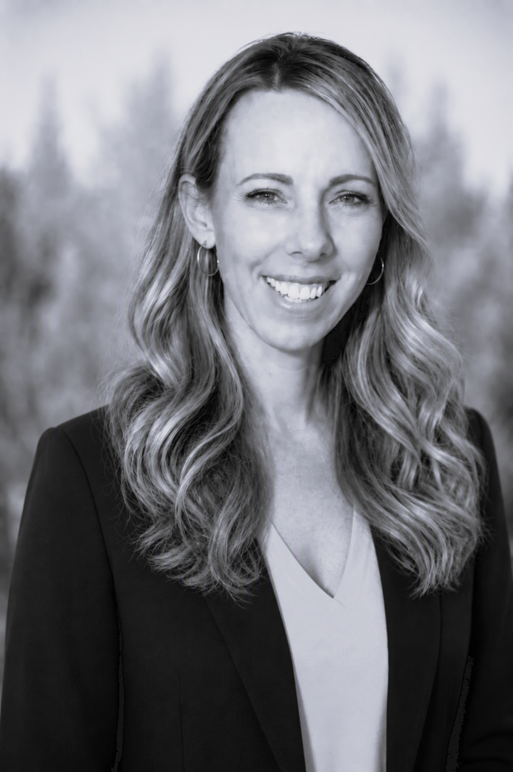 Black and white portrait of a smiling woman with long wavy hair, wearing a blazer and earrings, outdoors with blurred trees in the background.