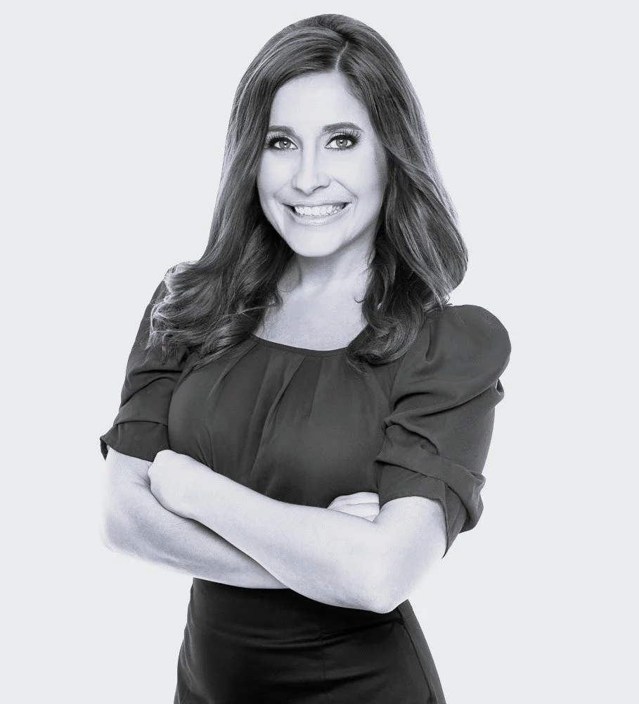 A woman with long, wavy hair smiling at the camera, standing with her arms crossed, wearing a dark blouse with puffed sleeves.