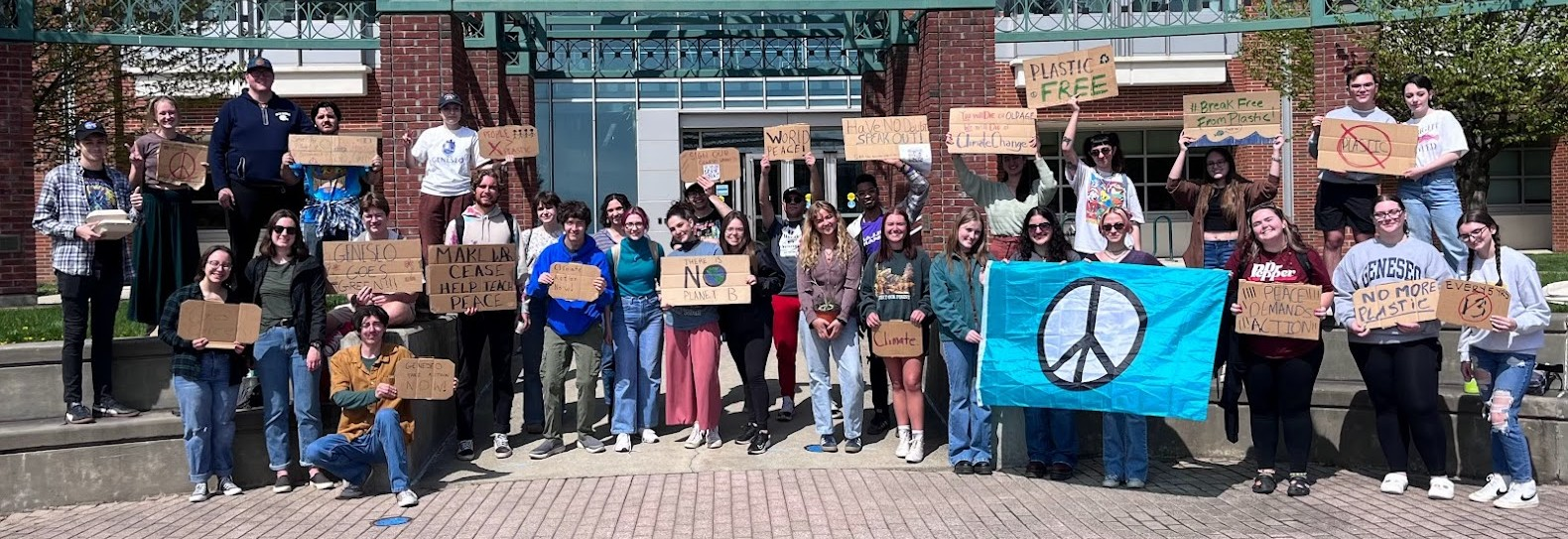 Group of people holding signs advocating for peace and environmental causes, standing outside a brick building. Some signs read "Plastic Free," "Make La Cease Help Tea Peace," and "No More Plastic." A person holds a blue peace flag.