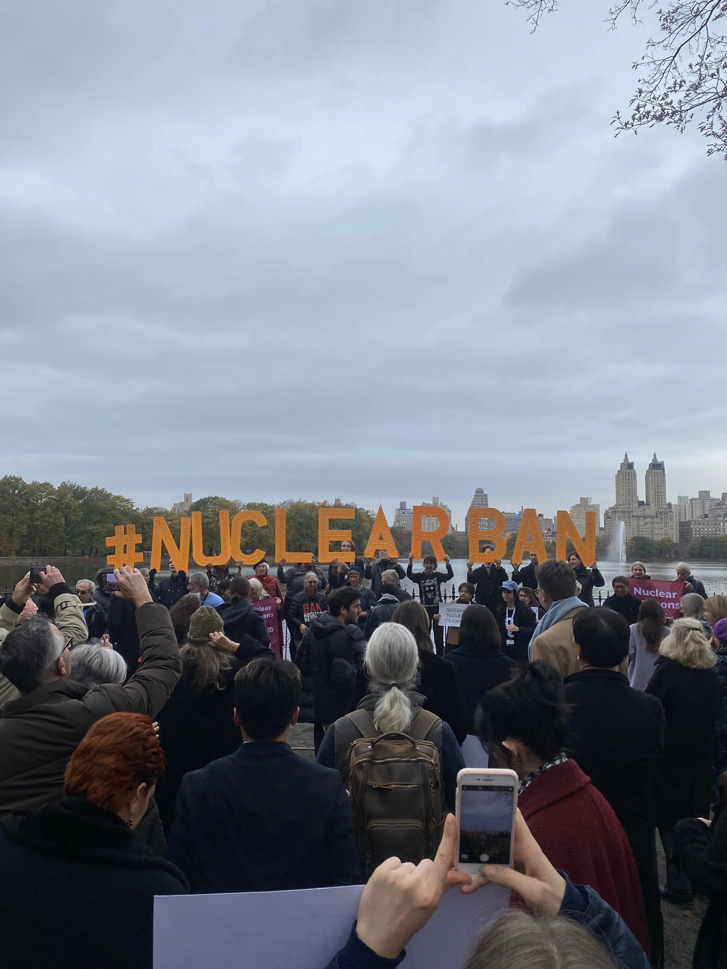 A crowd of people gathered outdoors holding a large orange sign that reads '#NUCLEAR BAN' with a city skyline, trees, and water in the background.