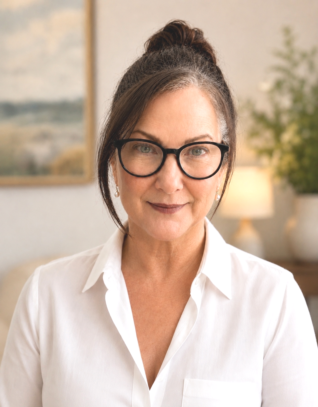 A middle-aged woman with glasses and a white shirt smiling in a cozy, well-lit room with artwork and a potted plant in the background.