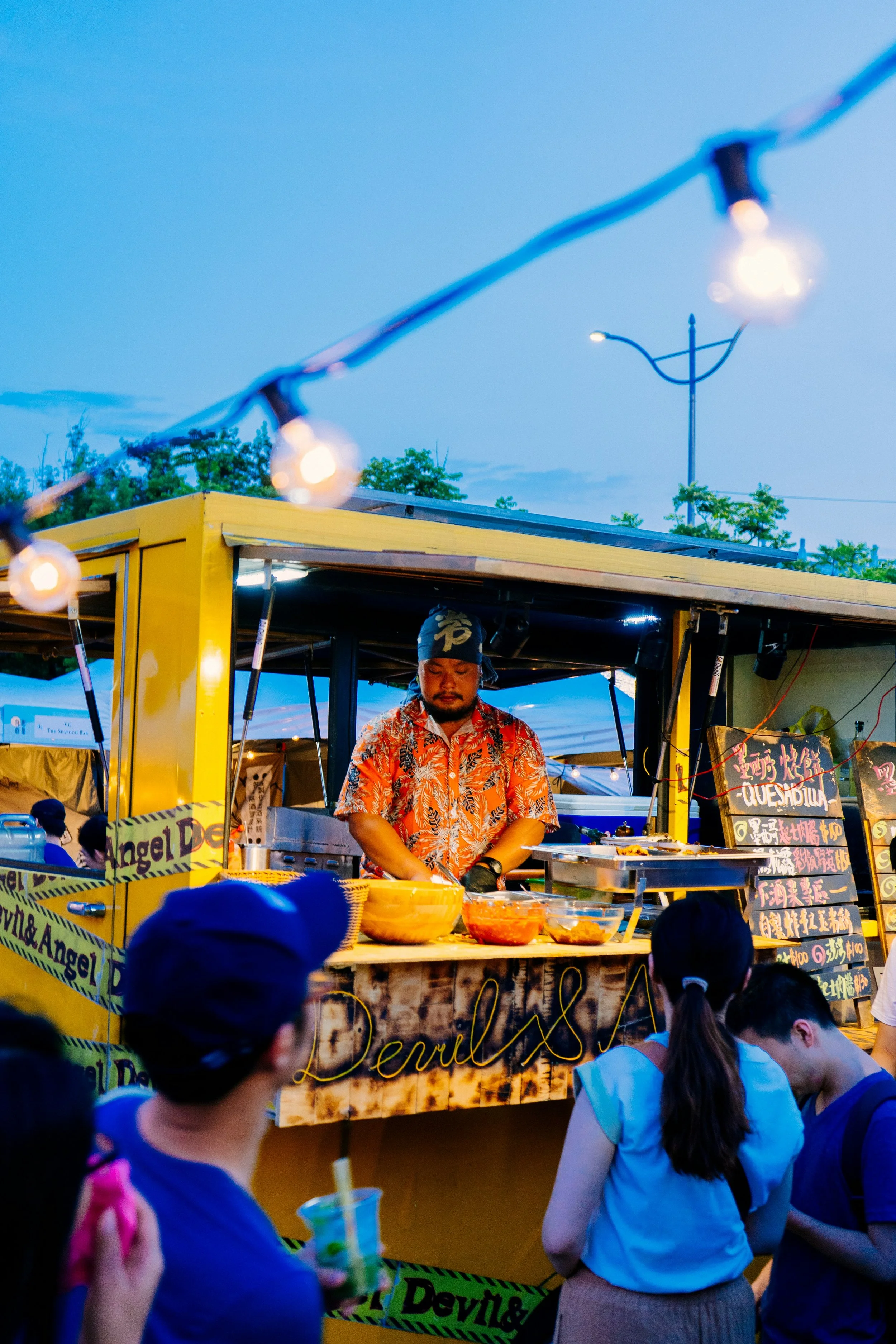A food stall at an outdoor night market, serving food to customers. The vendor is a man wearing a patterned shirt and a headscarf, with bowls of food in front of him. There are people in the crowd, some holding drinks. String lights decorate the scene and a sign with Chinese characters and the word 'Dewills' is visible.