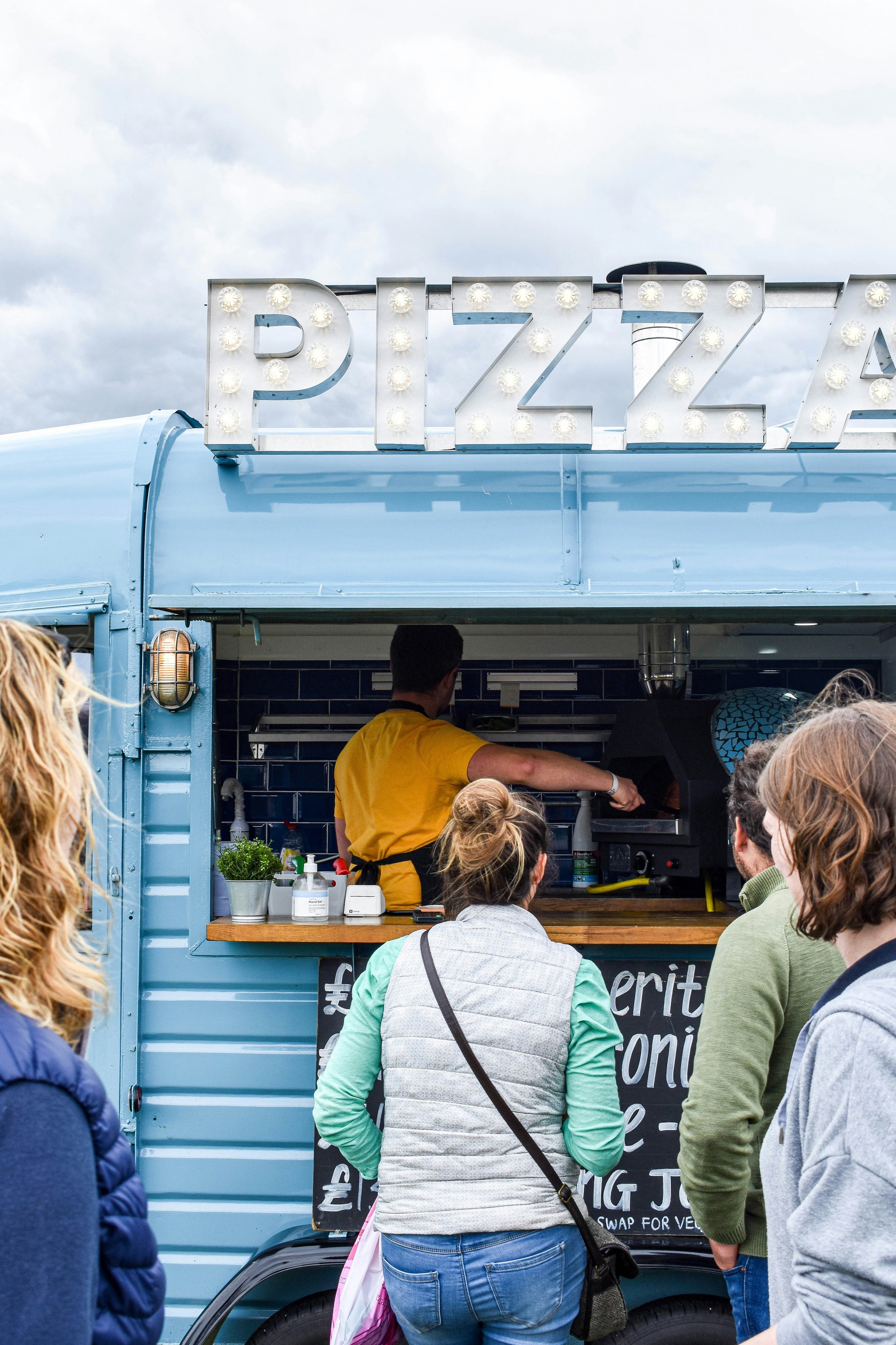 A blue food truck with a large illuminated sign that reads "PIZZA" on top. Several people are standing in line facing the truck, waiting to order.