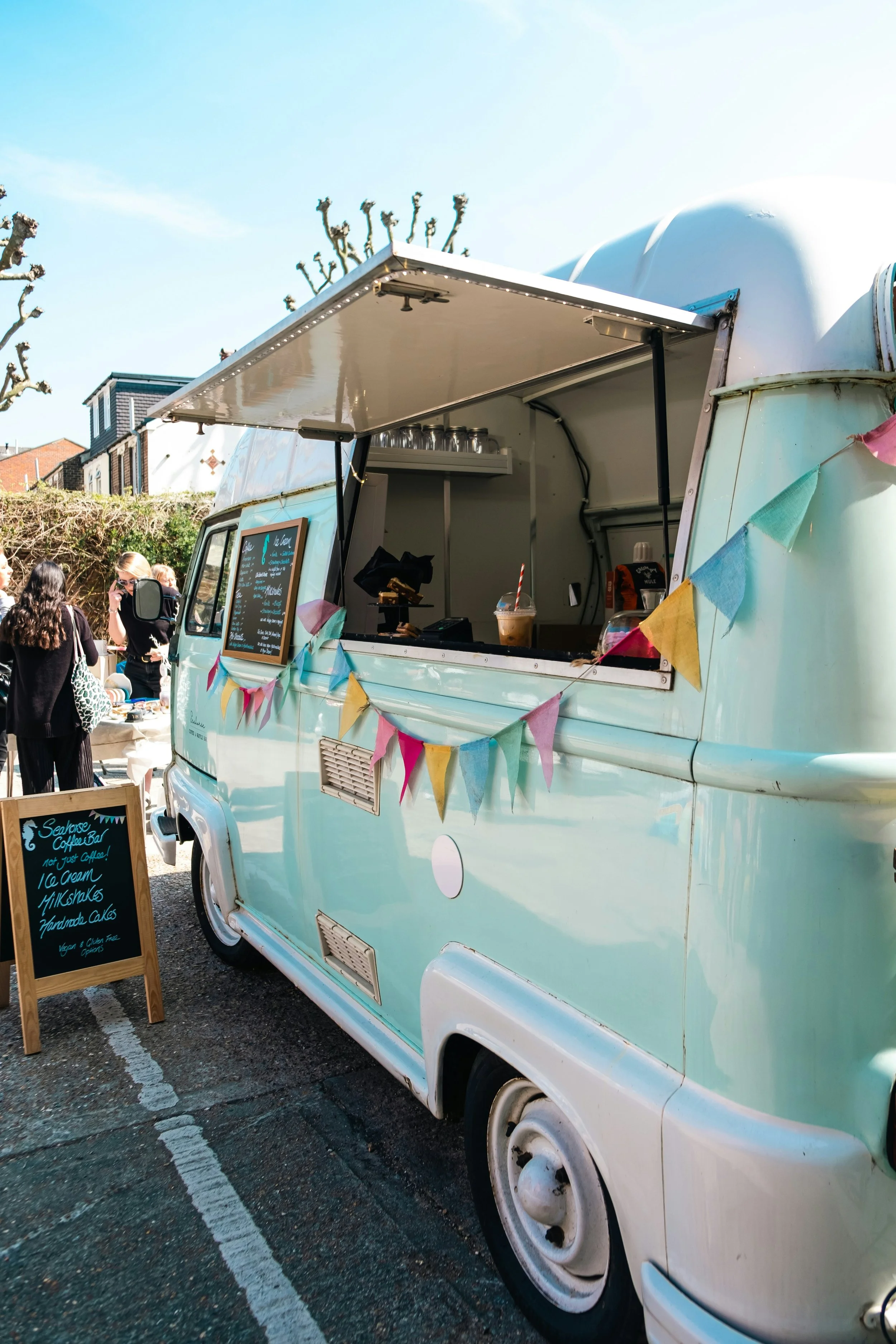 A light blue vintage food truck with colorful bunting, a menu board, and customers standing nearby on a sunny day.