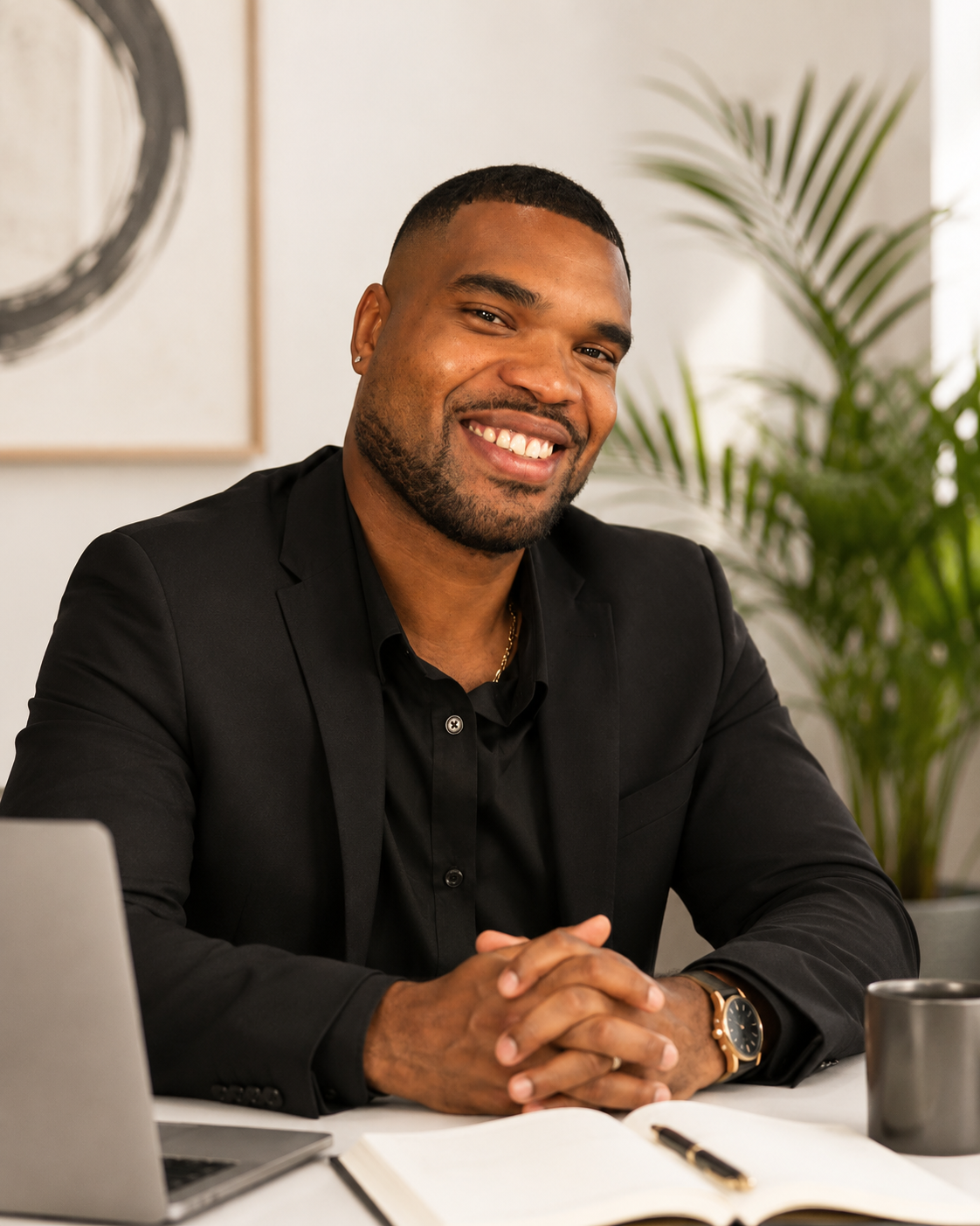 A smiling man in a black suit sitting at a desk with a laptop, notebook, pen, and mug, in a modern office with a large plant in the background.