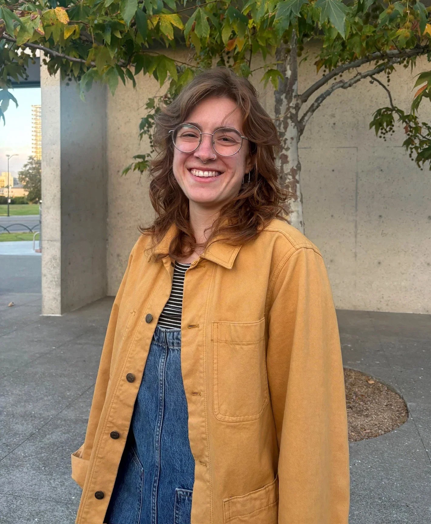 A young woman with shoulder-length wavy brown hair, glasses, and a septum piercing, smiling outdoors. She is wearing a yellow jacket over a black and white striped shirt, with a background of a beige wall, trees, and a cityscape in the distance.