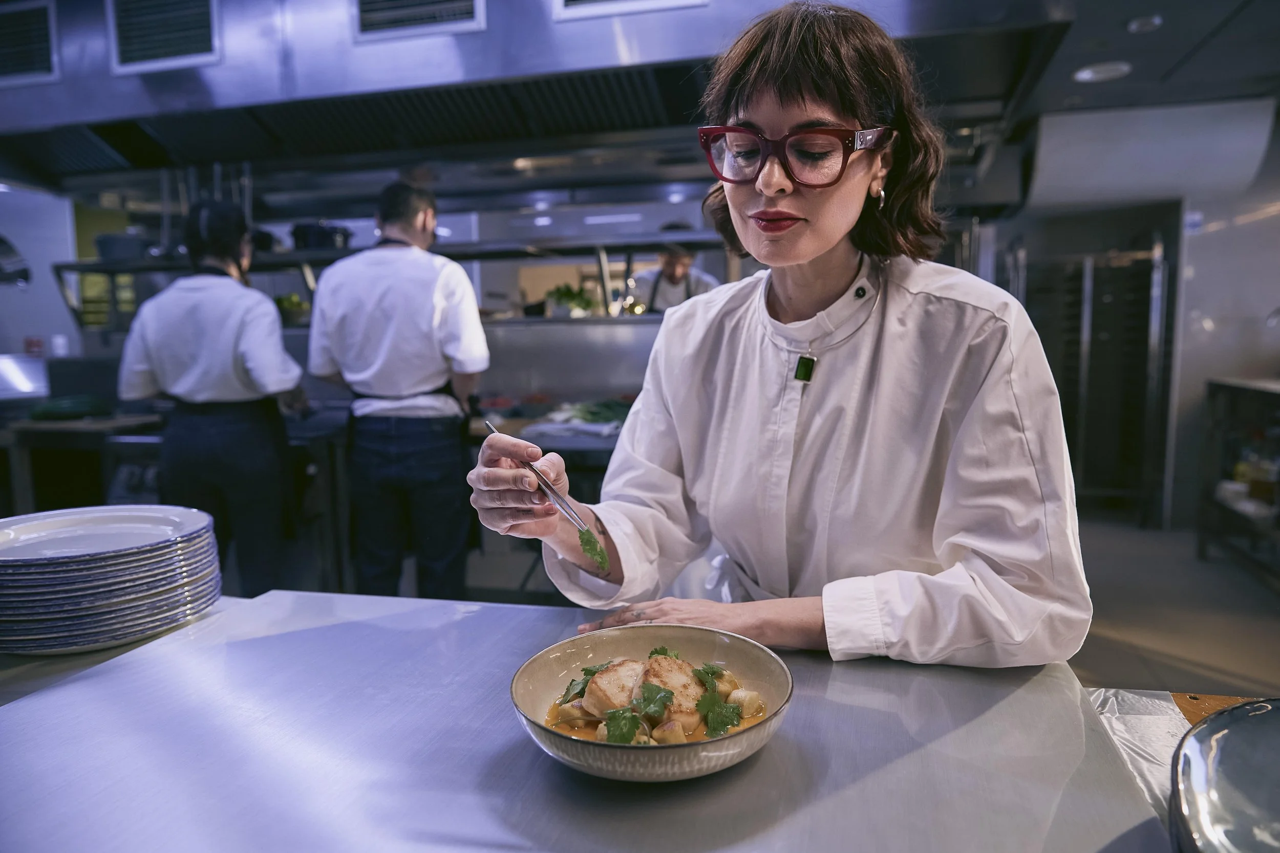 Una chef con gafas rojas y cabello castaño oscuro prepara un plato en una cocina profesional, con otros cocineros en el fondo.