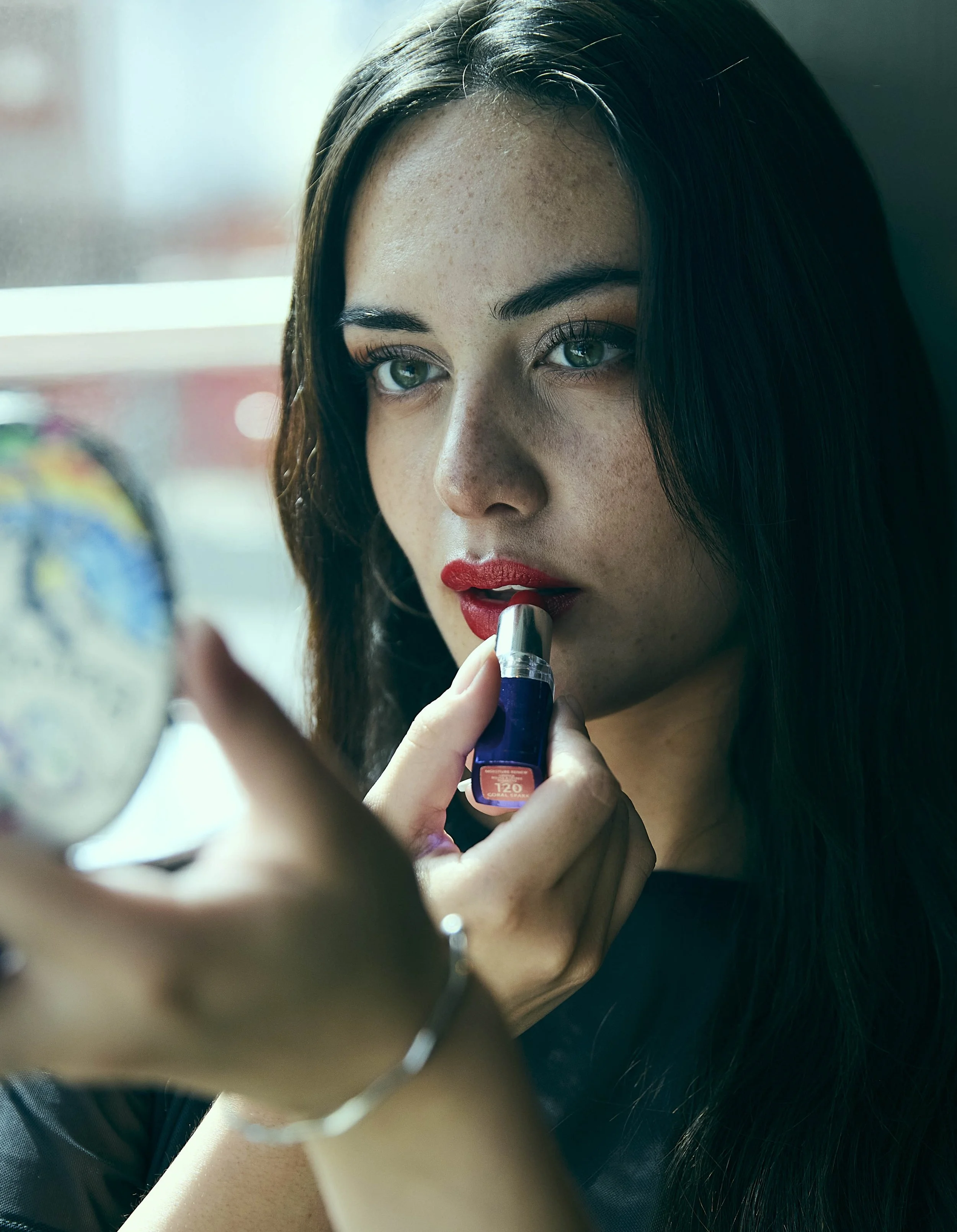 Mujer con cabello castaño, ojos azules y labios rojos, aplicándose lápiz labial mientras se mira en un espejo.