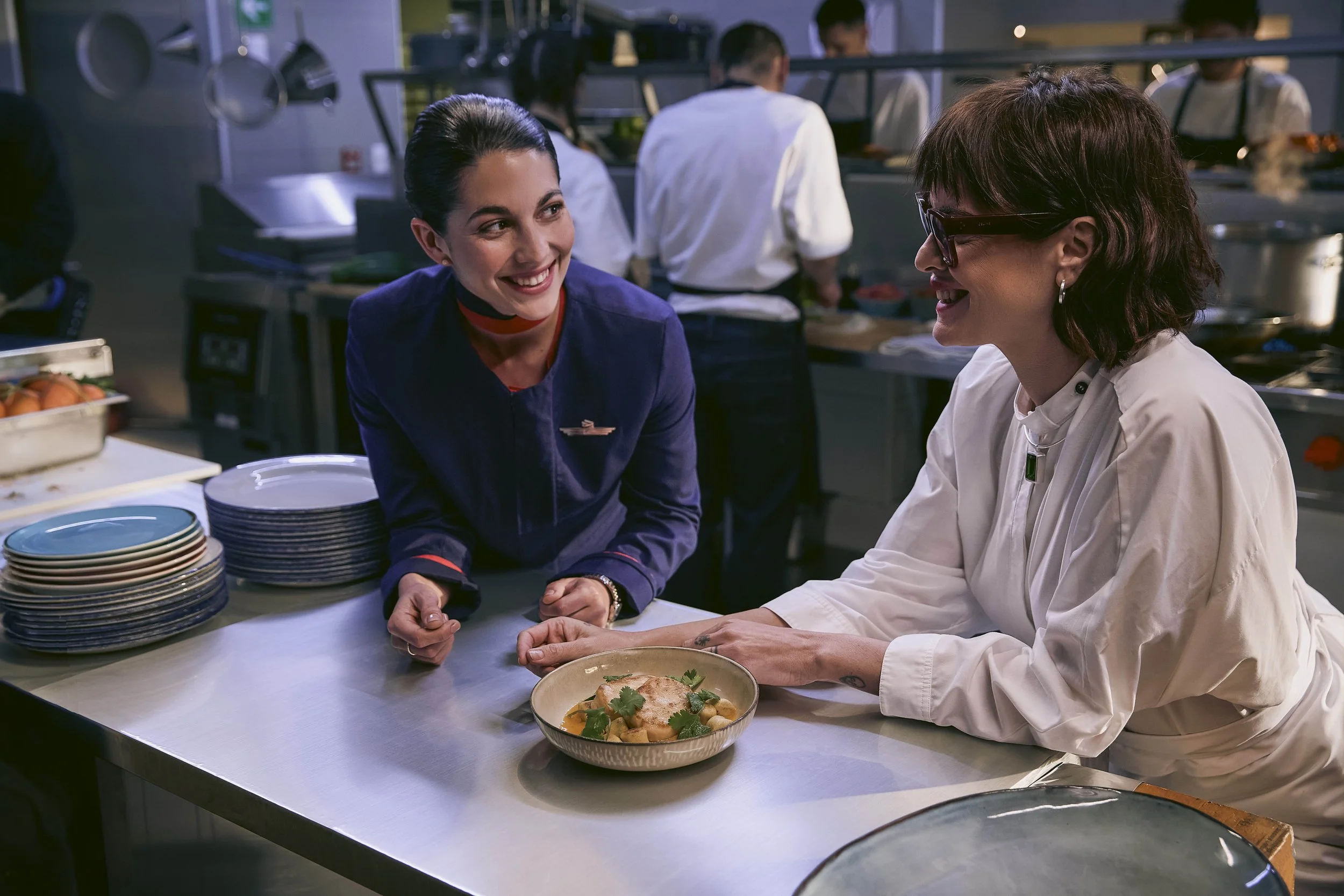 Dos mujeres conversando y sonriendo en una cocina de restaurante, con un plato de comida en la mesa y personal de cocina en el fondo.