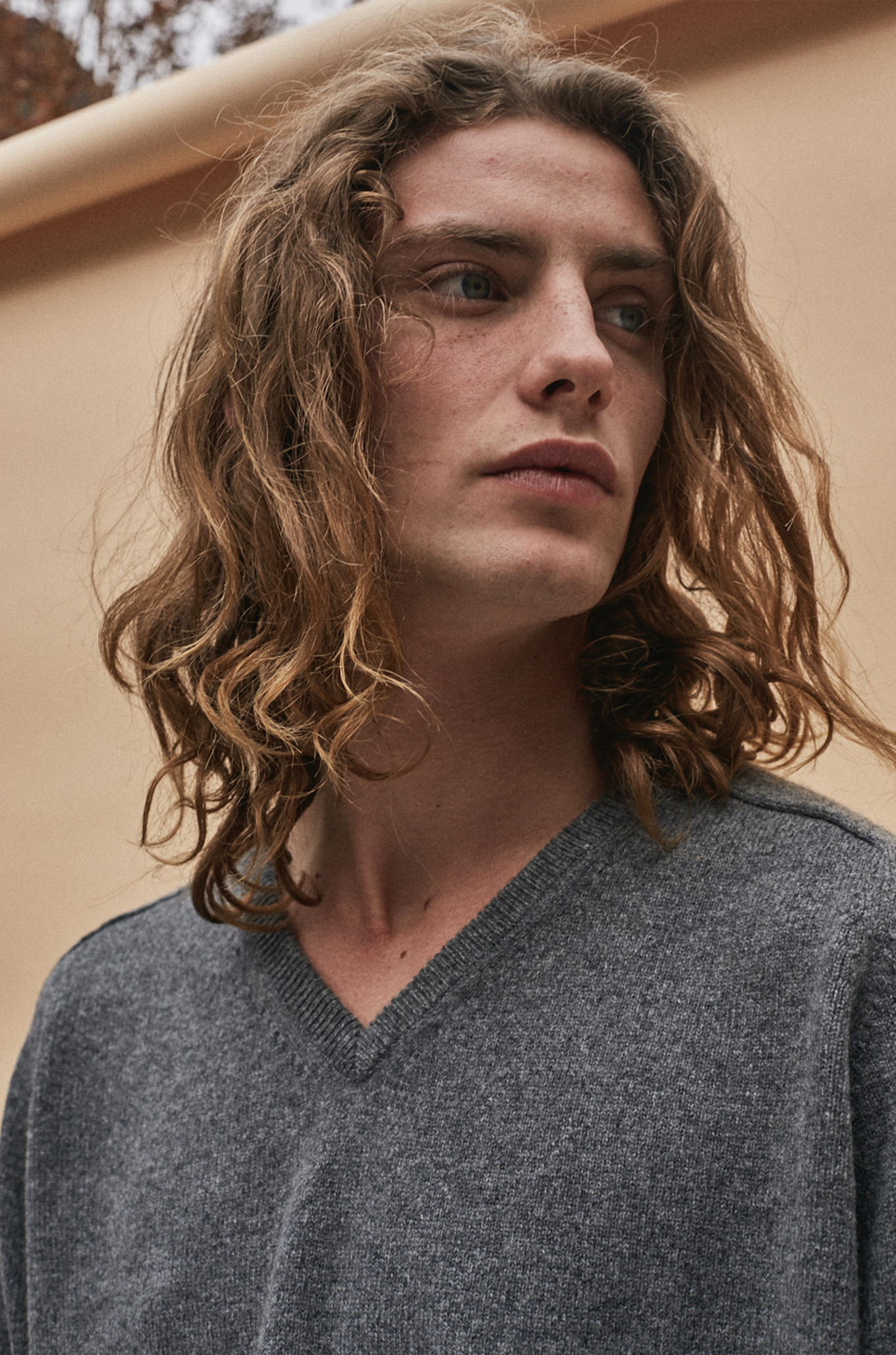 Joven con cabello rizado, rubio y largos, usando suéter gris, con expresión pensativa.