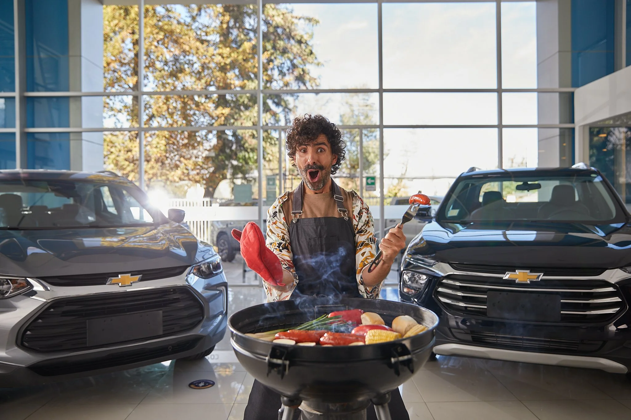 Un hombre feliz con barba y cabello rizado, usando delantal, sosteniendo un tenedor y una manzana, en un concesionario de autos con carros Chevrolet y ventanas grandes con vista a árboles de otoño.