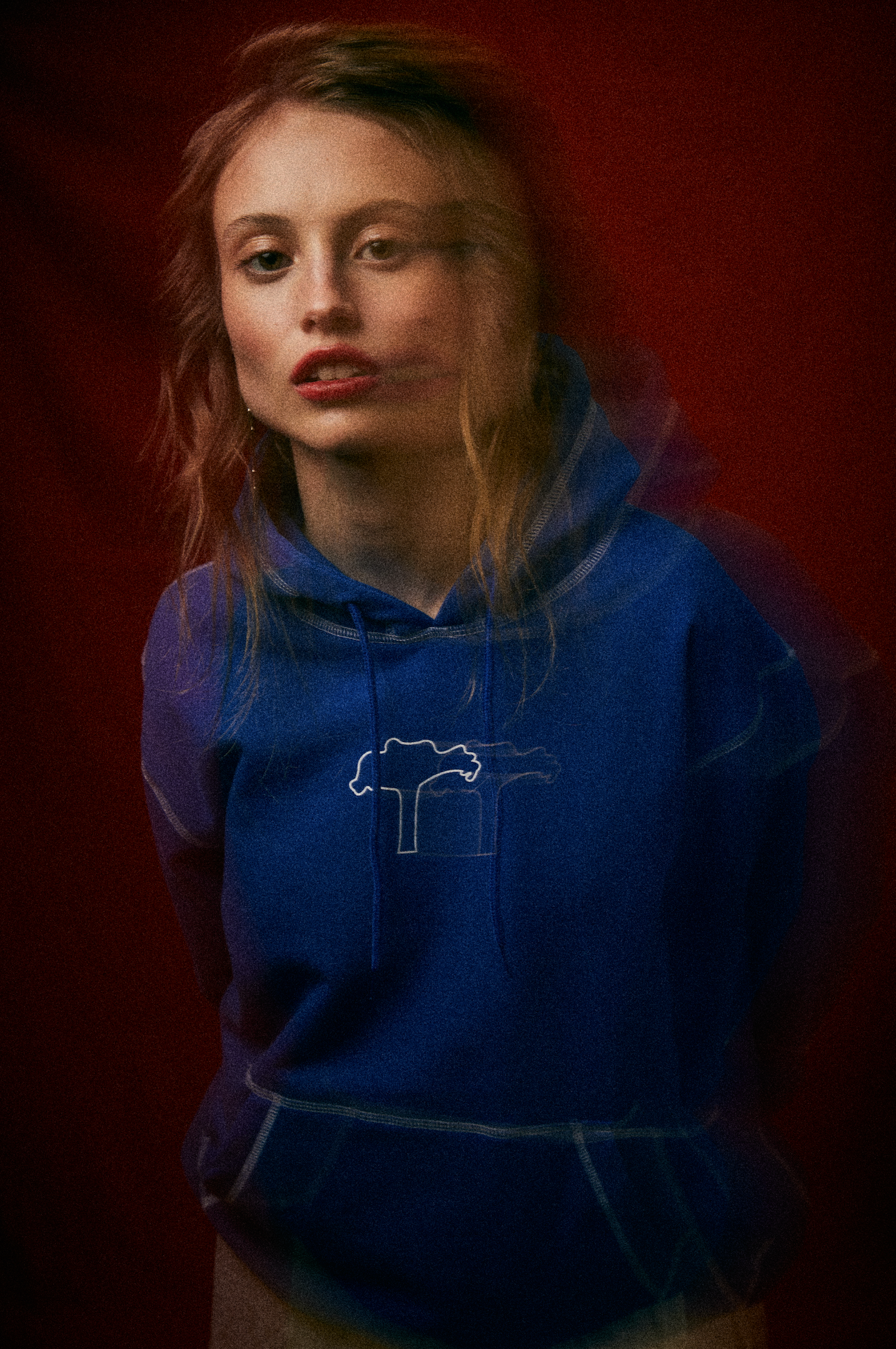Joven mujer con cabello rizado, usando sudadera azul con un dibujo de un árbol, en un fondo rojo oscuro.