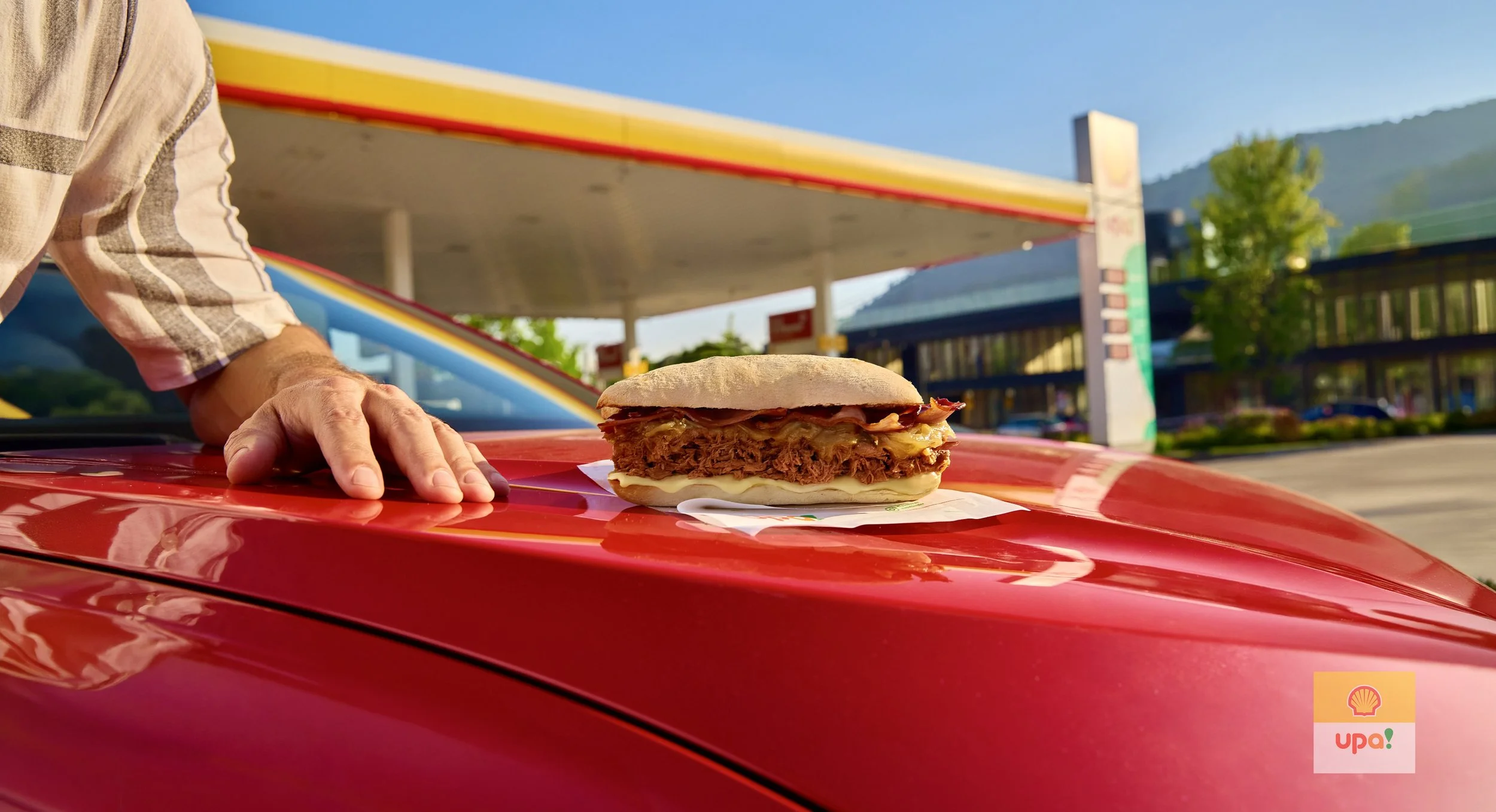 Primer plano de una hamburguesa en un coche rojo frente a una gasolinera en un día soleado, con un fondo de montañas verdes.