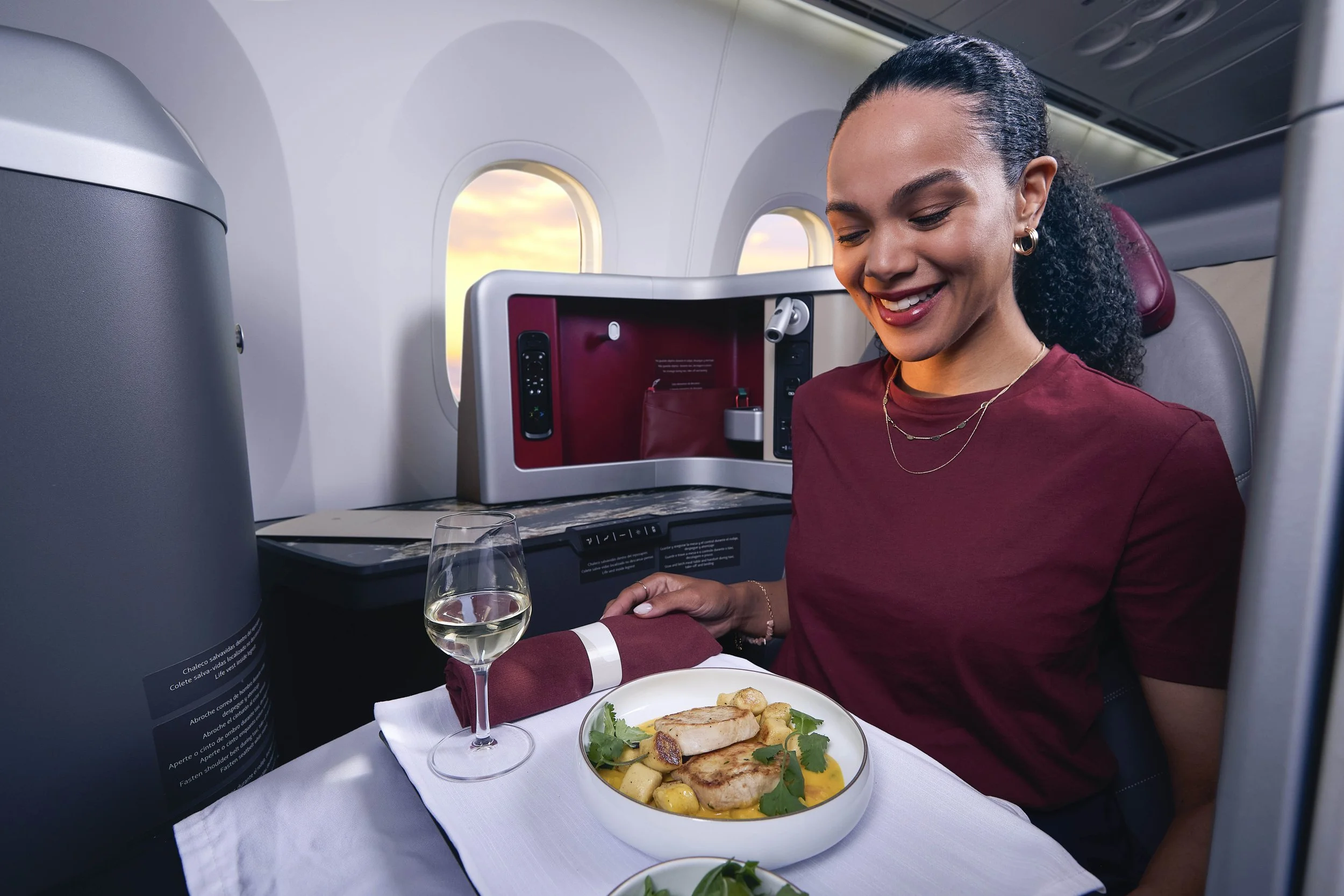 Mujer sonriendo en un avión con cena en la mesa, vista de la ventana mostrando un atardecer.