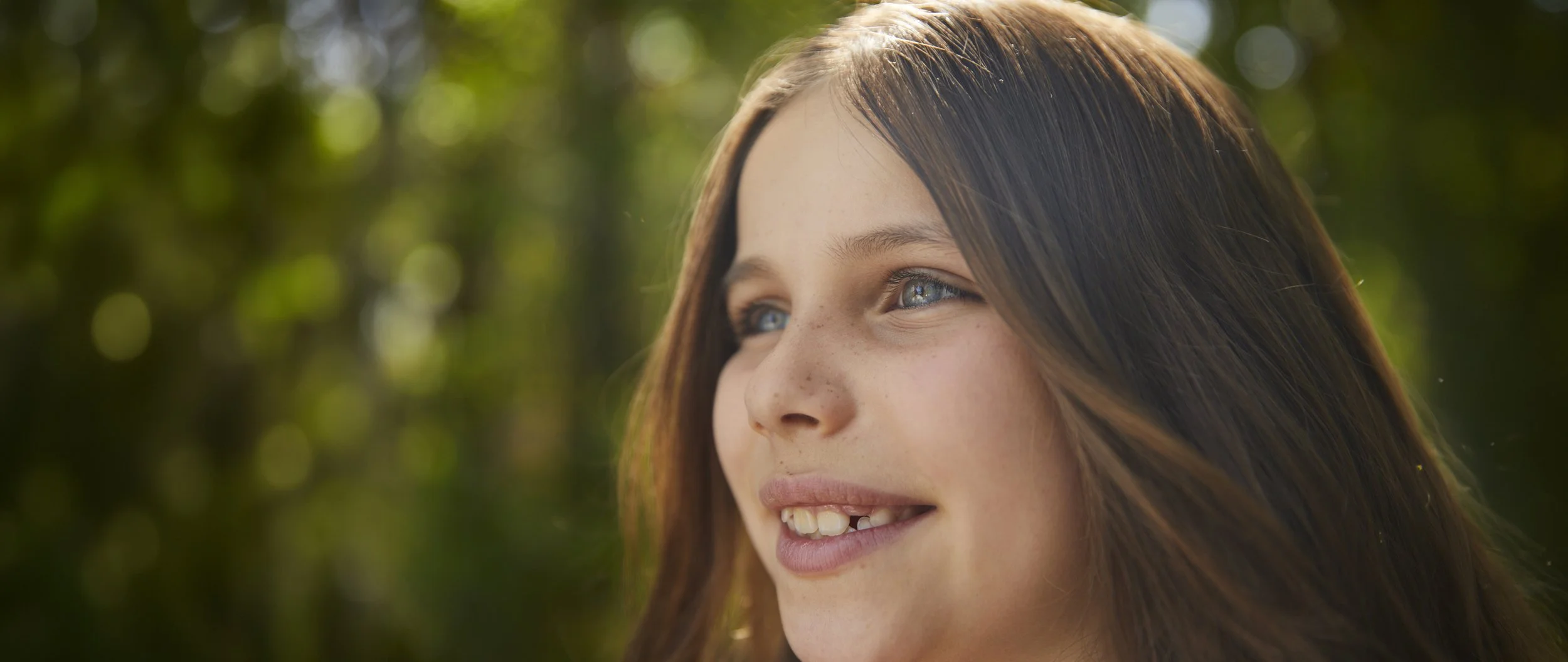 Retrato de una joven con cabello castaño, ojos azules y sonrisa, en un entorno natural con fondo desenfocado de árboles y luz solar.