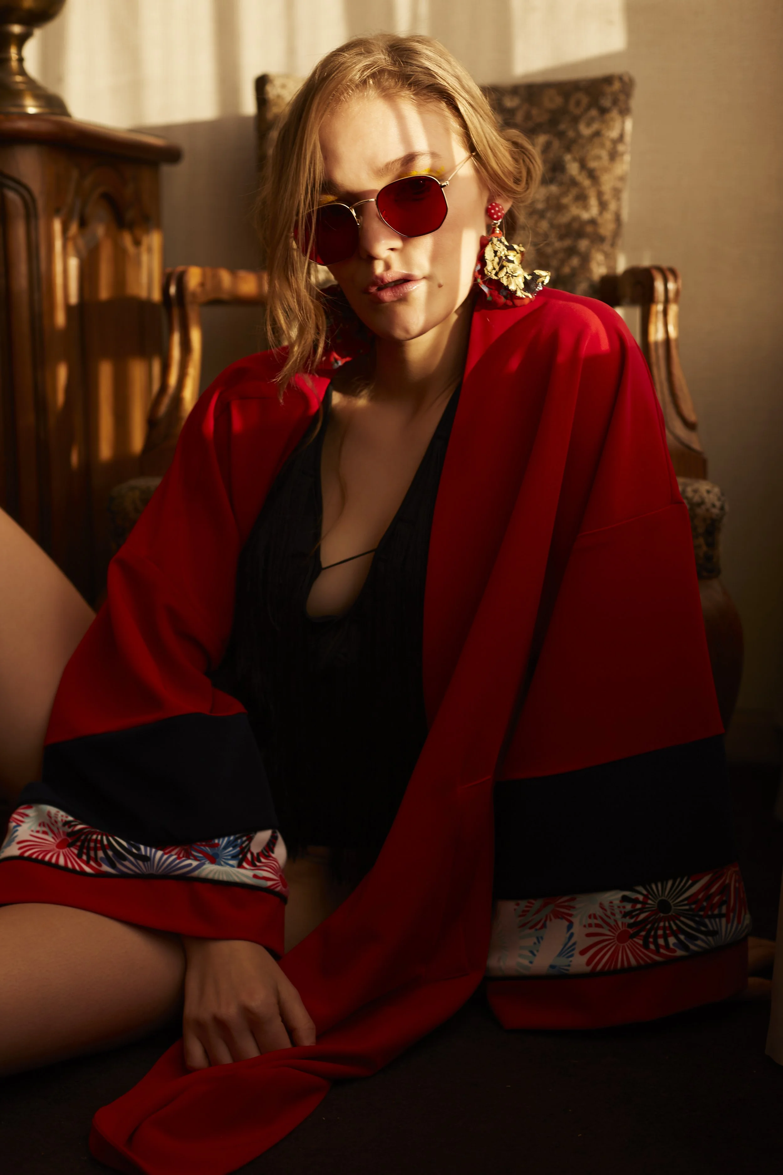 Mujer con gafas de sol redondas de cristales oscuros, large aro dorado, y aretes grandes con detalles dorados y rojos, vestida con un kimono rojo y ropa interior negra, sentada en una silla de madera con tapizado floral y sosteniendo una falda o pren