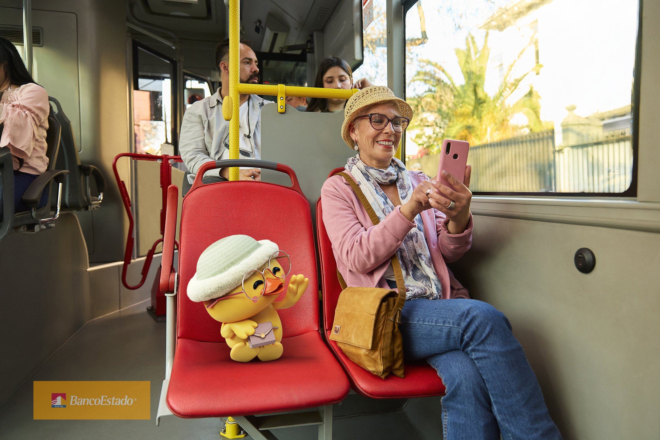 Una mujer de la tercera edad con gafas, sombrero de paja, bufanda, camisa rosa y bolso marrón, sonriendo y usando su teléfono móvil en un autobús. Frente a ella, en la primera fila, hay una figura de una pata de goma con gafas, bufanda, sombrero de p