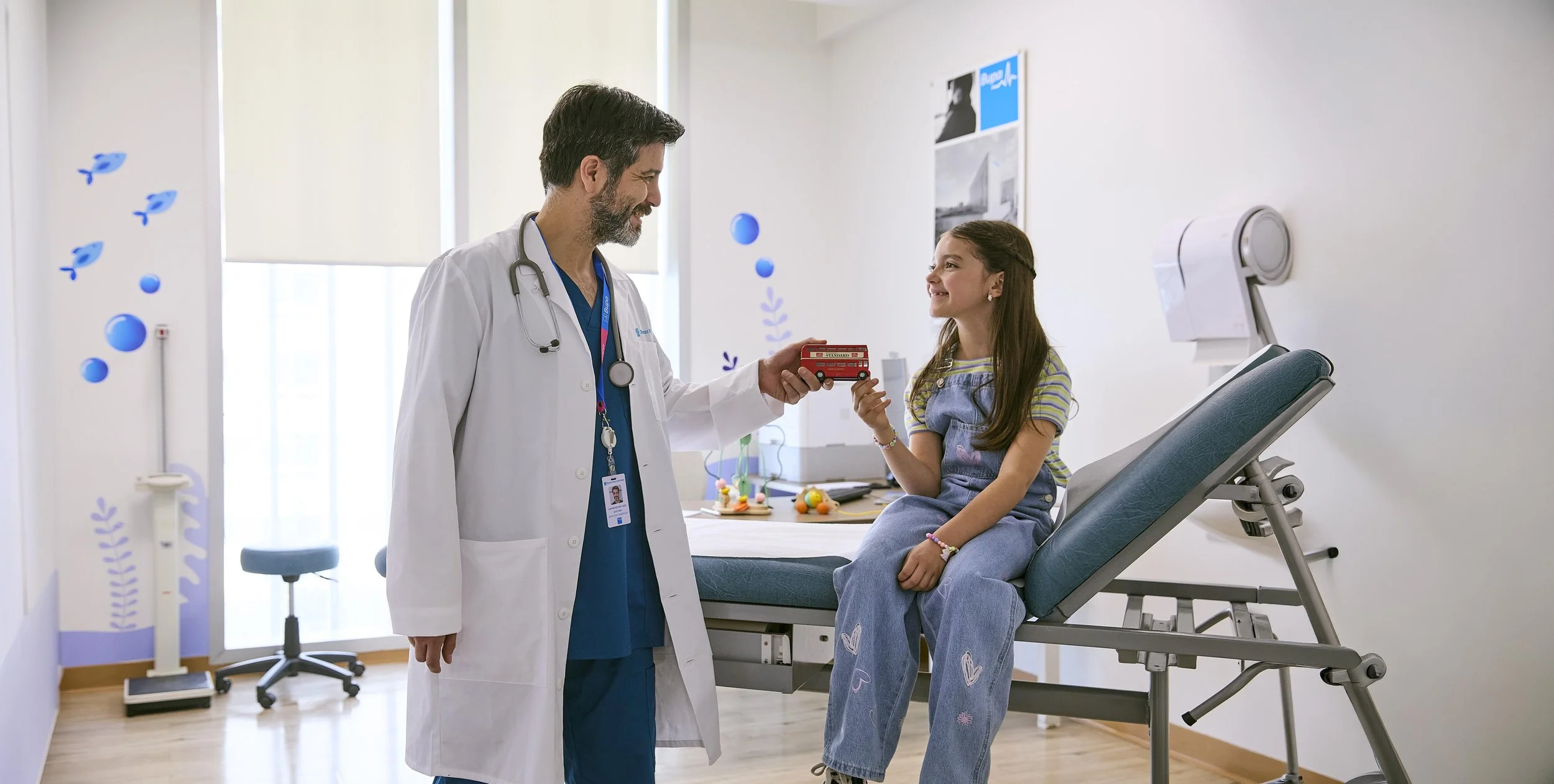 Médico y niña en consulta en un hospital infantil, el médico le entrega un juguete de autobús rojo a la niña que está sentada en una camilla, la niña sonríe.