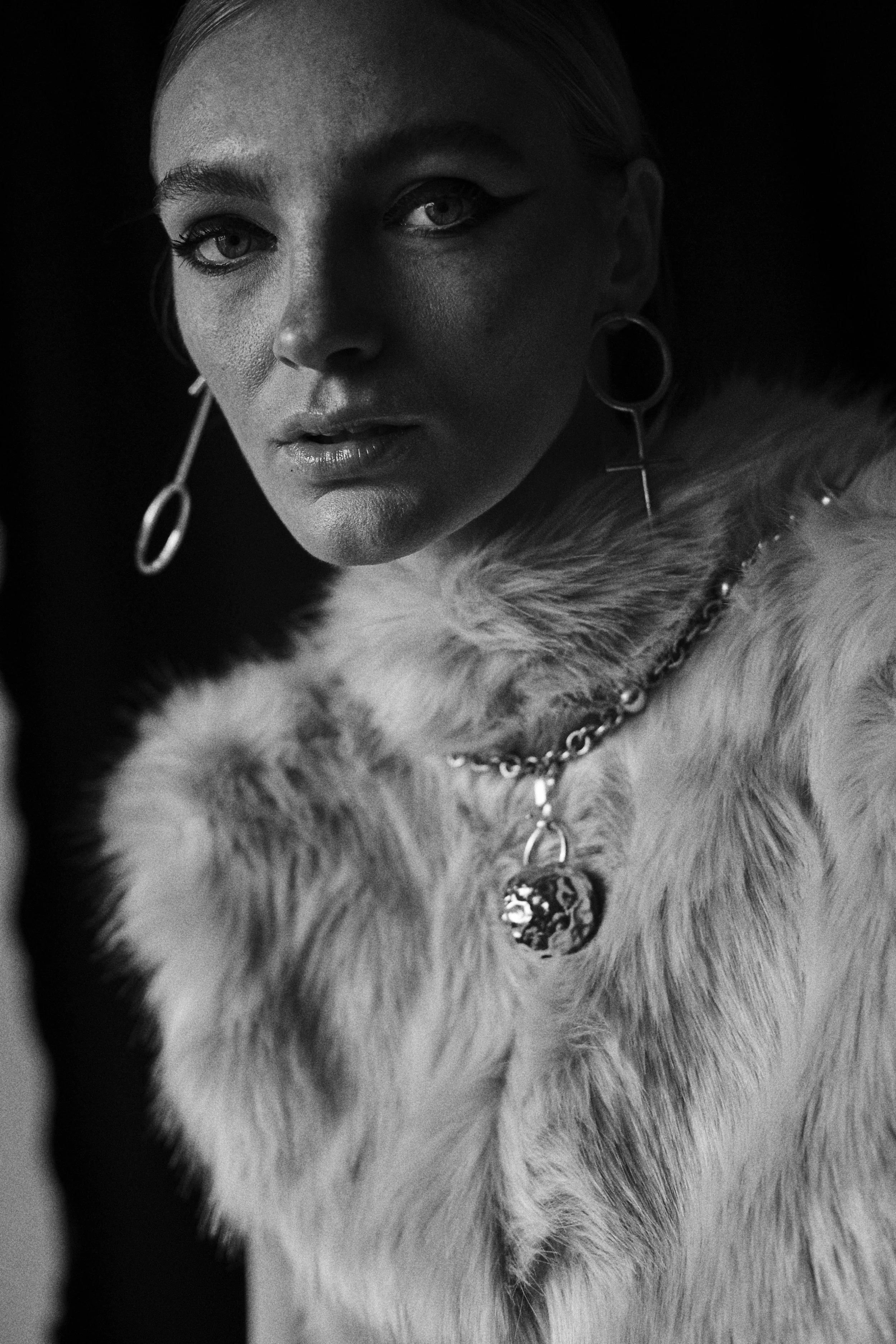 Retrato de una mujer con pendientes grandes, collar con colgante, luciendo un abrigo de piel y maquillaje elaborado, en un estilo artístico en blanco y negro.