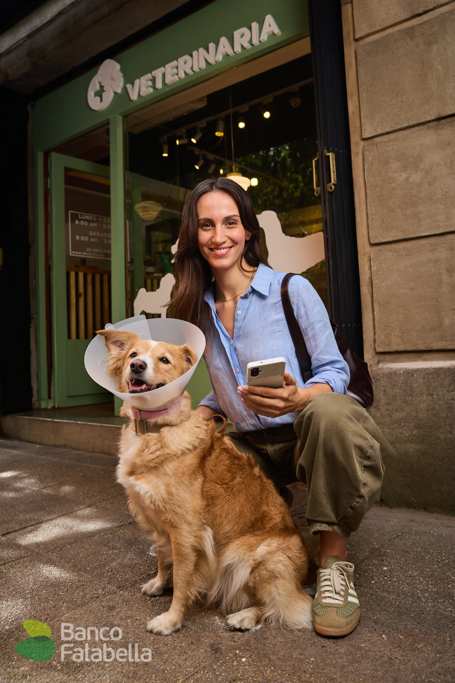 Mujer sonriente con un perro con collar ortopédico afuera de una veterinaria llamada Banco Falabella. La mujer sostiene un teléfono móvil y usa ropa casual. La veterinaria tiene un cartel con información en la puerta.