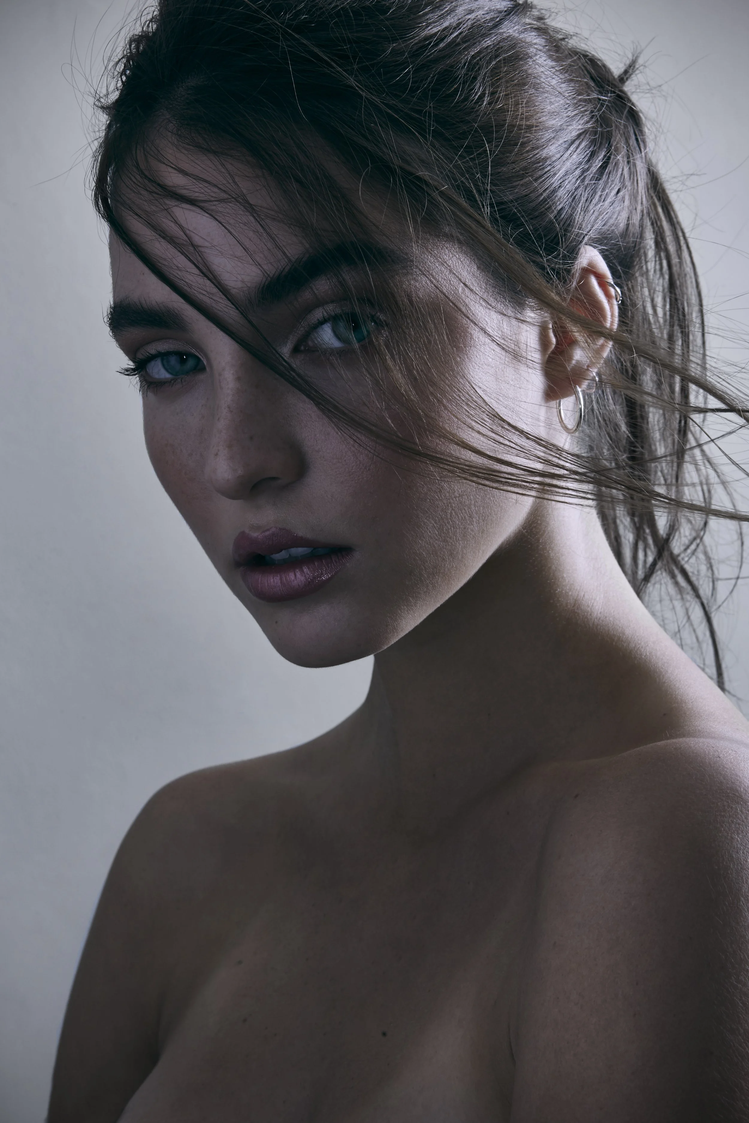 Primer plano de una mujer con cabello castaño oscuro, ojos azules, piel con pecas, usando pendientes de aro, fotografiada en luz natural y fondo neutro.