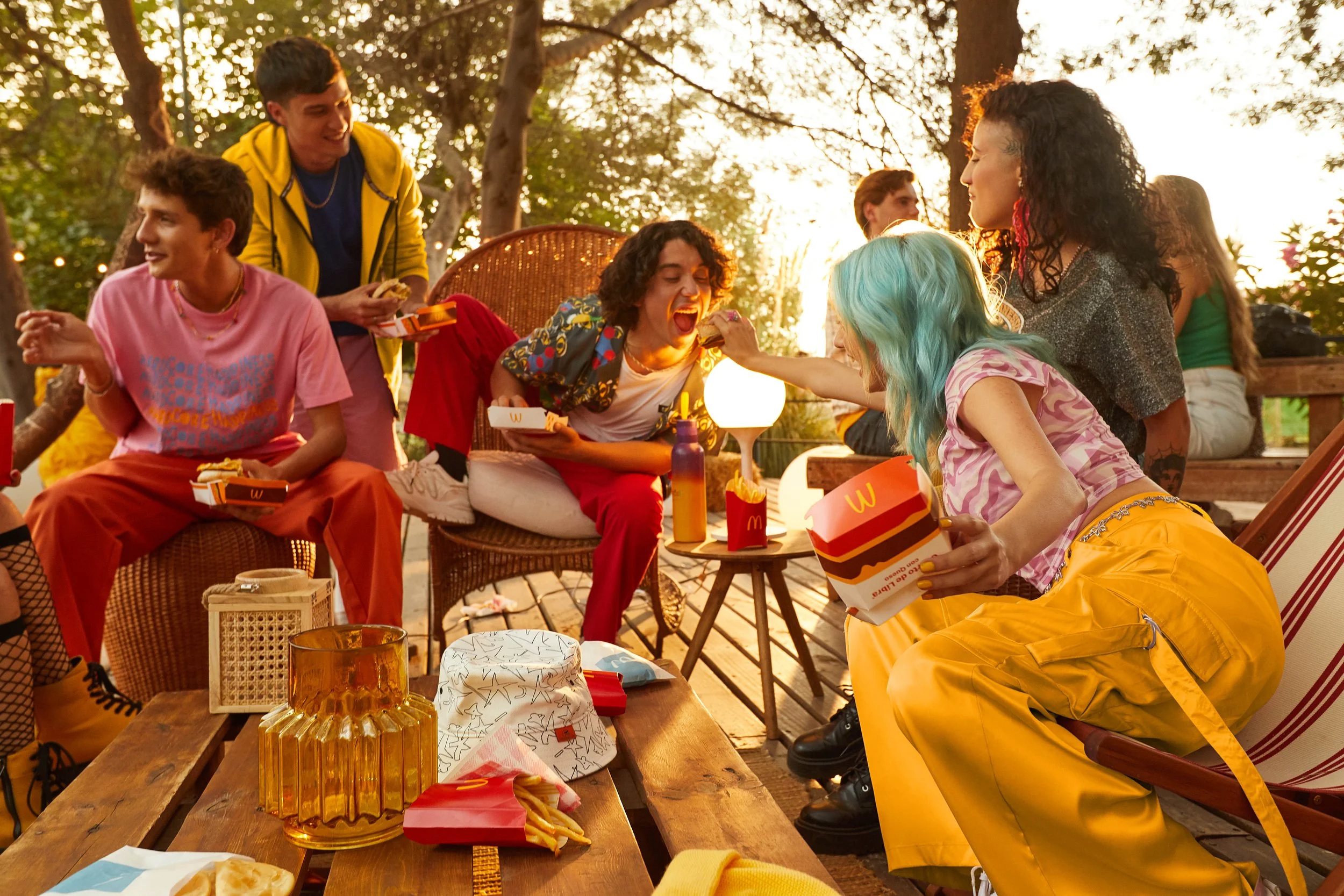Grupo de jóvenes disfrutando una comida con comida rápida en un ambiente al aire libre durante el atardecer, rodeados de árboles y luz cálida.