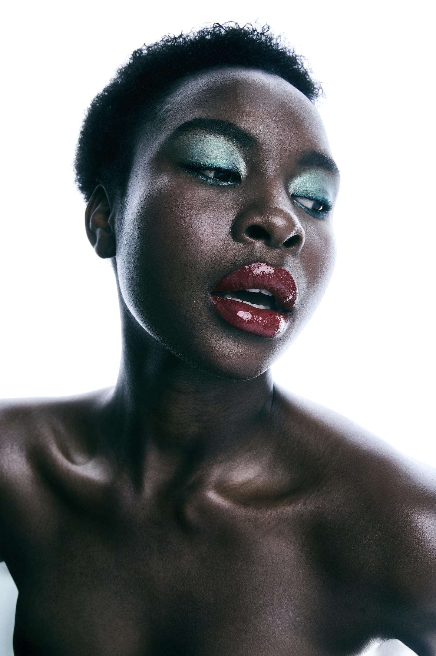 Retrato de una mujer de piel oscura con maquillaje en tonos verdes y labios rojos, mostrando la parte superior del cuerpo y el rostro en primer plano con fondo blanco.