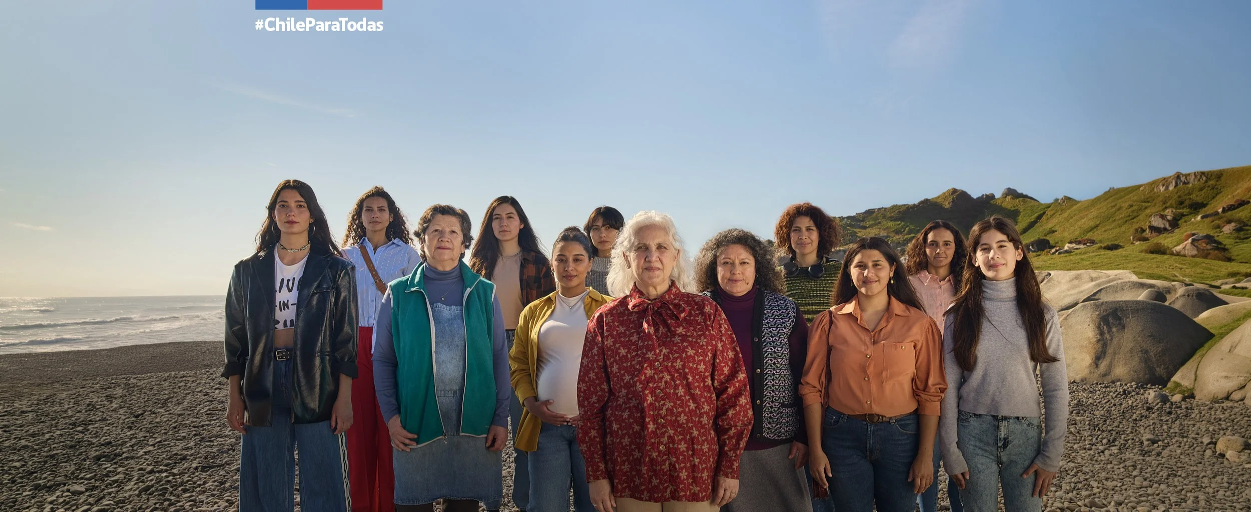 Grupo diverso de mujeres jóvenes y mayores en una playa con colinas verdes al fondo, bajo un cielo despejado.