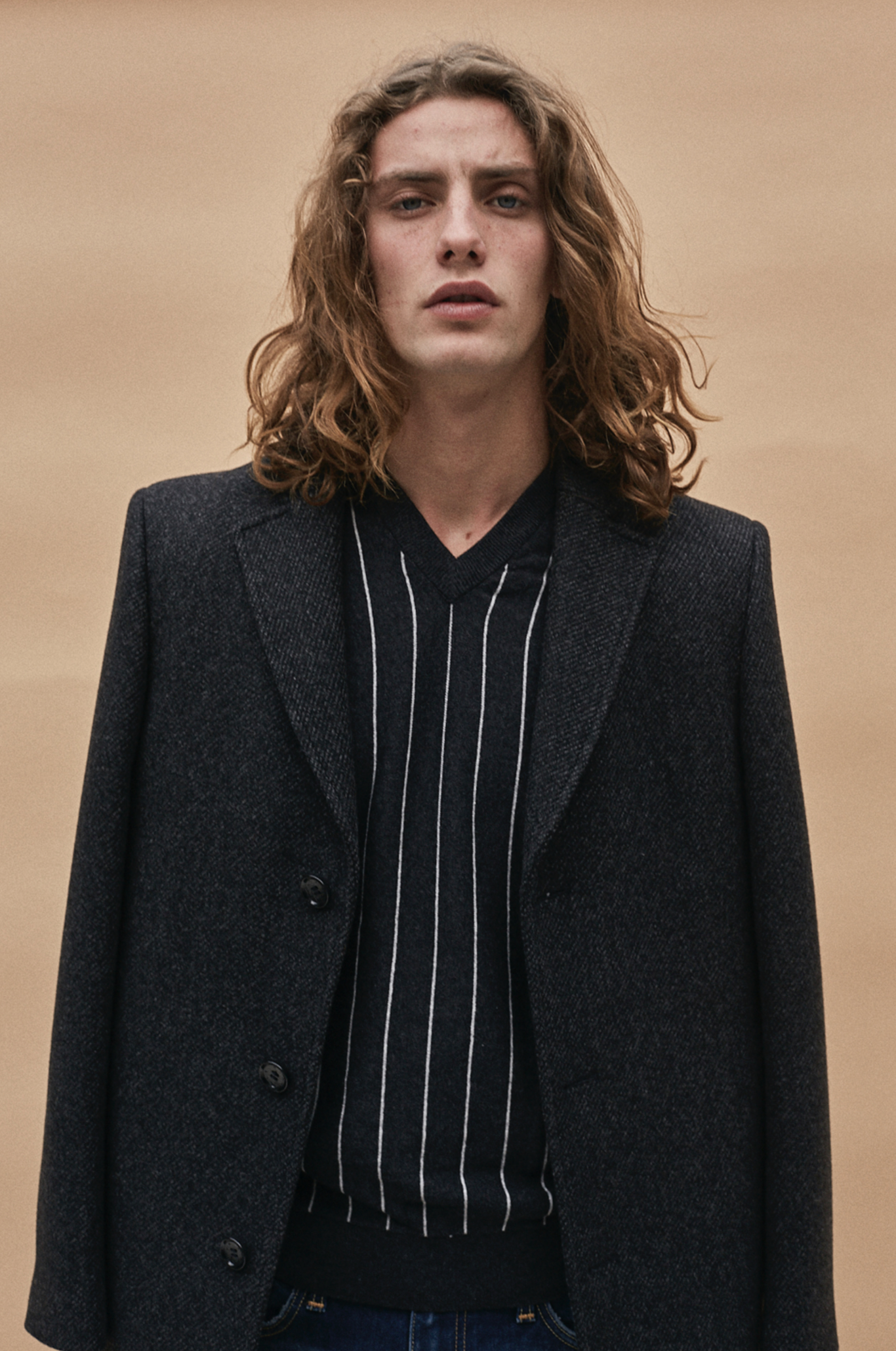 Joven con cabello rizado y expresión facial seria, vistiendo chaqueta oscura y suéter a rayas, de pie contra un fondo beige.