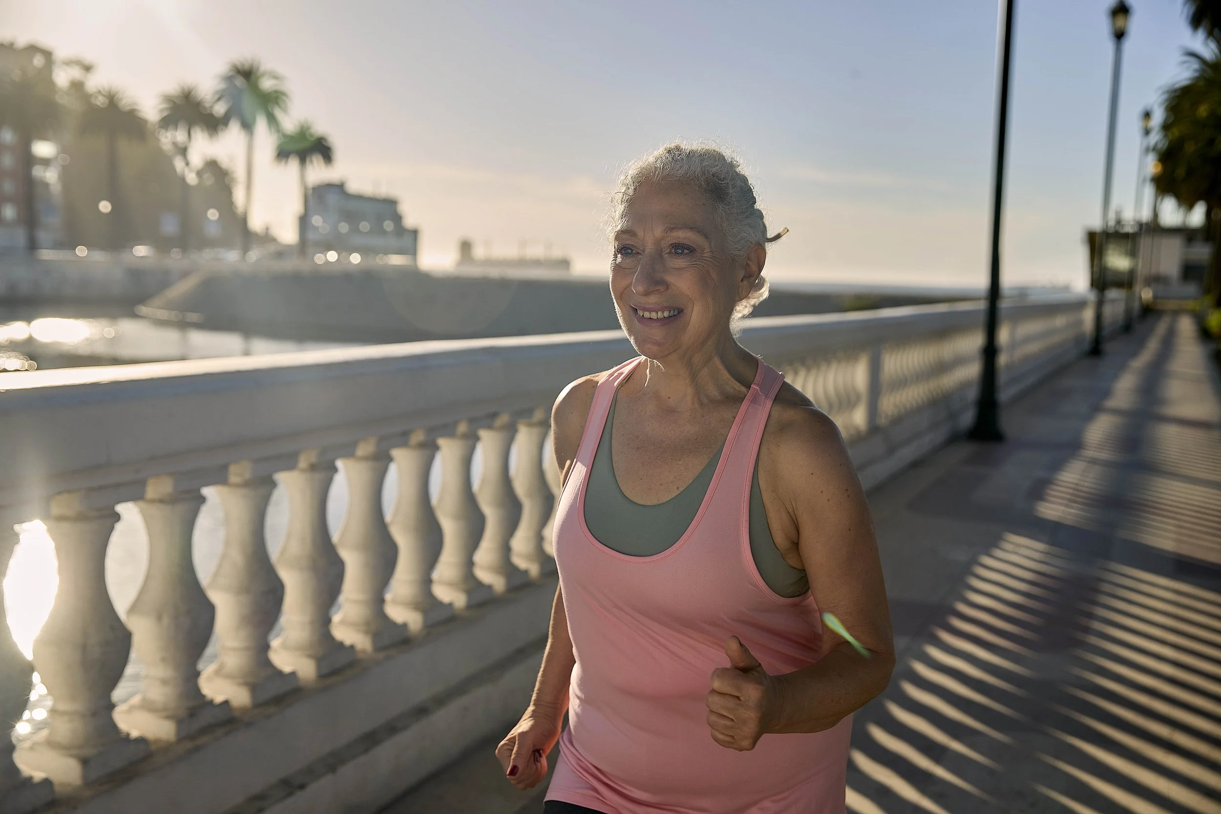 Mujer anciana corriendo al aire libre en un paseo junto al mar con palmeras y farolas, durante el atardecer.