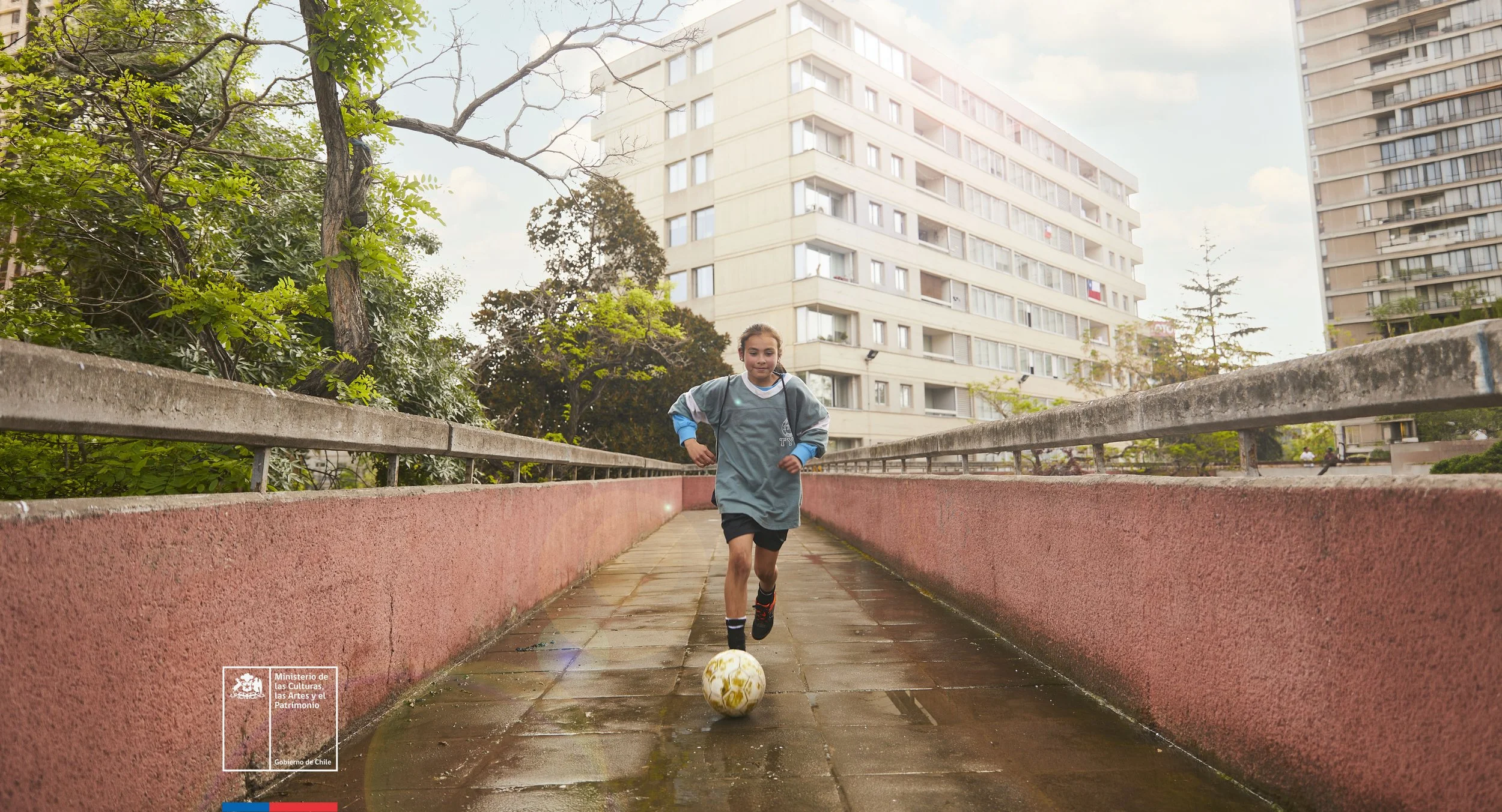 Una niña corriendo en una calle peatonal con una pelota de fútbol. Está vestida con ropa deportiva y lleva auriculares. Al fondo hay edificios altos y árboles, el cielo está parcialmente nublado.