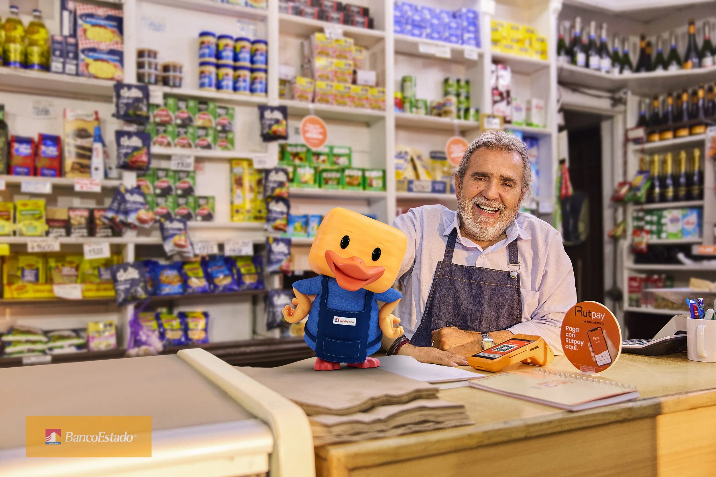 Un hombre mayor en una tienda, vestido con delantal y sonriendo, acompañado de un muñeco con rostro de pato. La tienda tiene estanterías llenas de productos, y en el mostrador hay un lector de tarjetas y un cartel naranja que invita a pagar con Rutpa