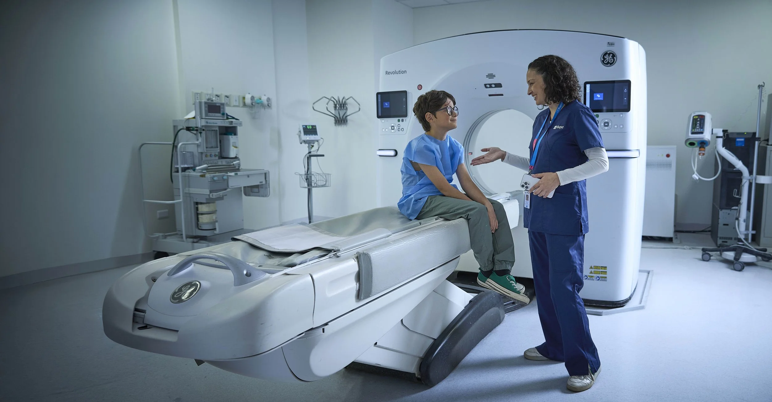 Una enfermera está conversando con una joven paciente en una sala de radiología con una máquina de tomografía. La paciente está sentada en la camilla de la máquina y la enfermera la está explicando.