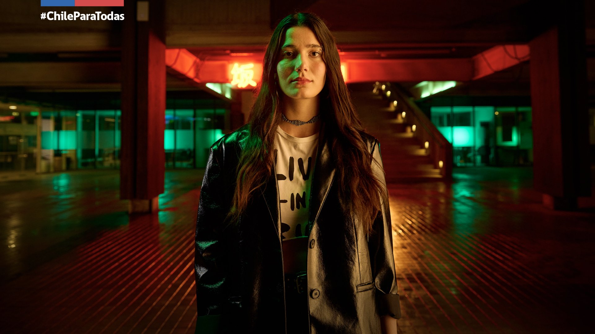 Joven mujer con cabello largo y suelto, vestida con chaqueta de cuero y camiseta blanca con letras, de pie en un lugar con luces de neón verdes y rojas, y escaleras de fondo.