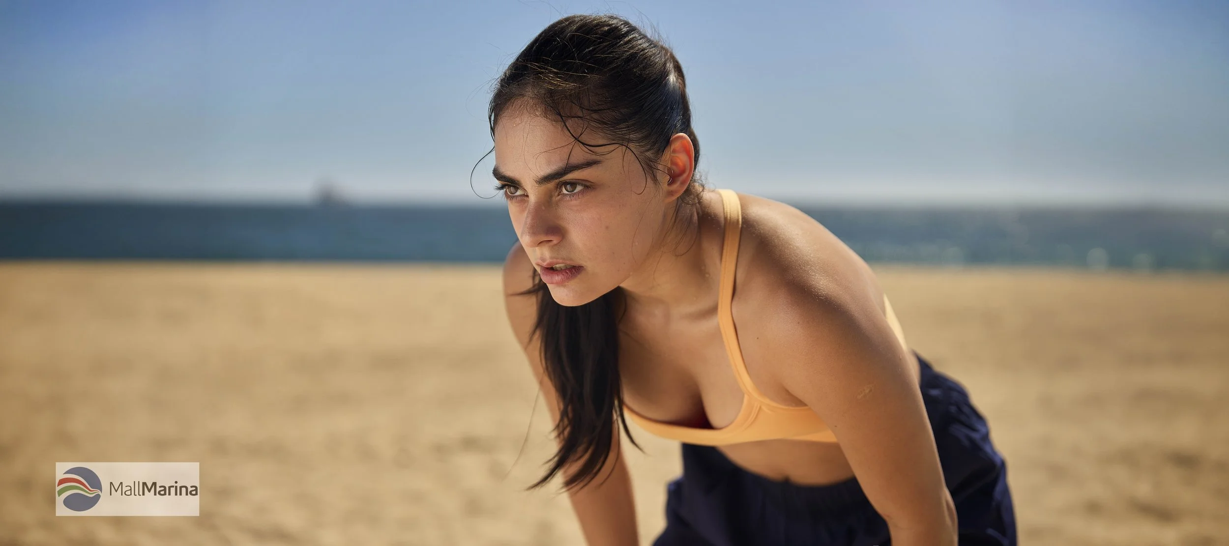 Una mujer joven con cabello oscuro y piel clara, usando un top naranja y pantalones deportivos oscuros, está en una playa, inclinada hacia adelante, con una expresión de concentración o esfuerzo.
