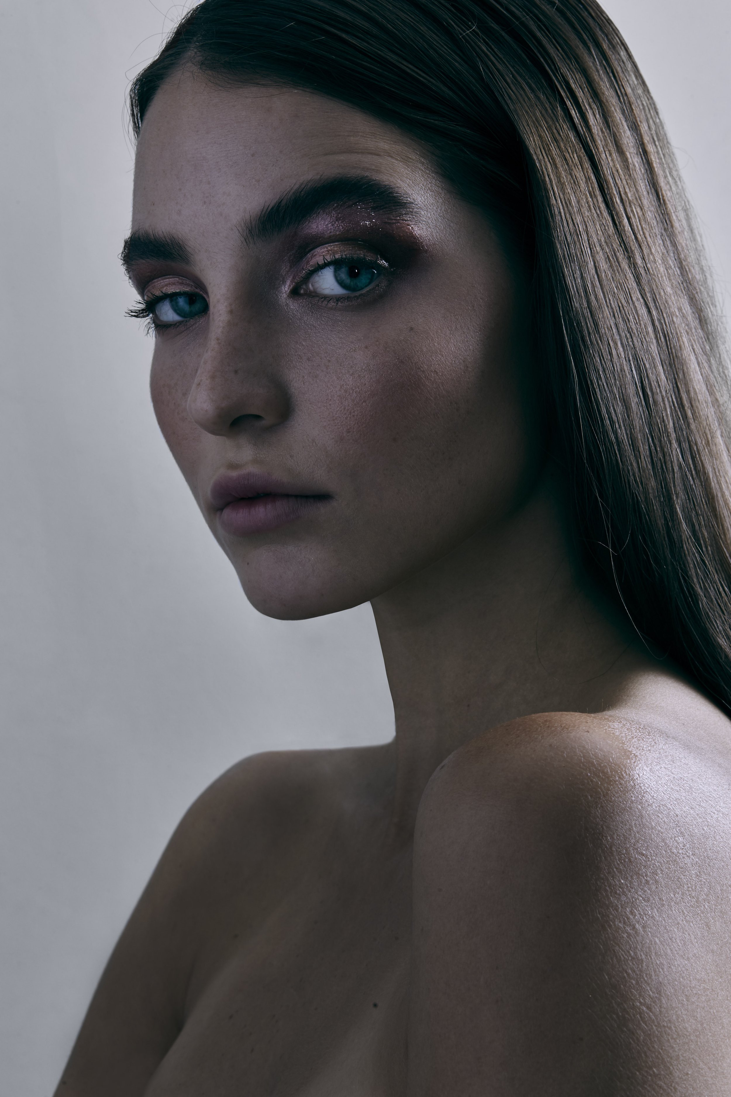 Retrato de una mujer con cabello largo y rubio, piel con pecas, ojos azules y maquillaje de ojos en tonos rosa y morado, con expresión seria.