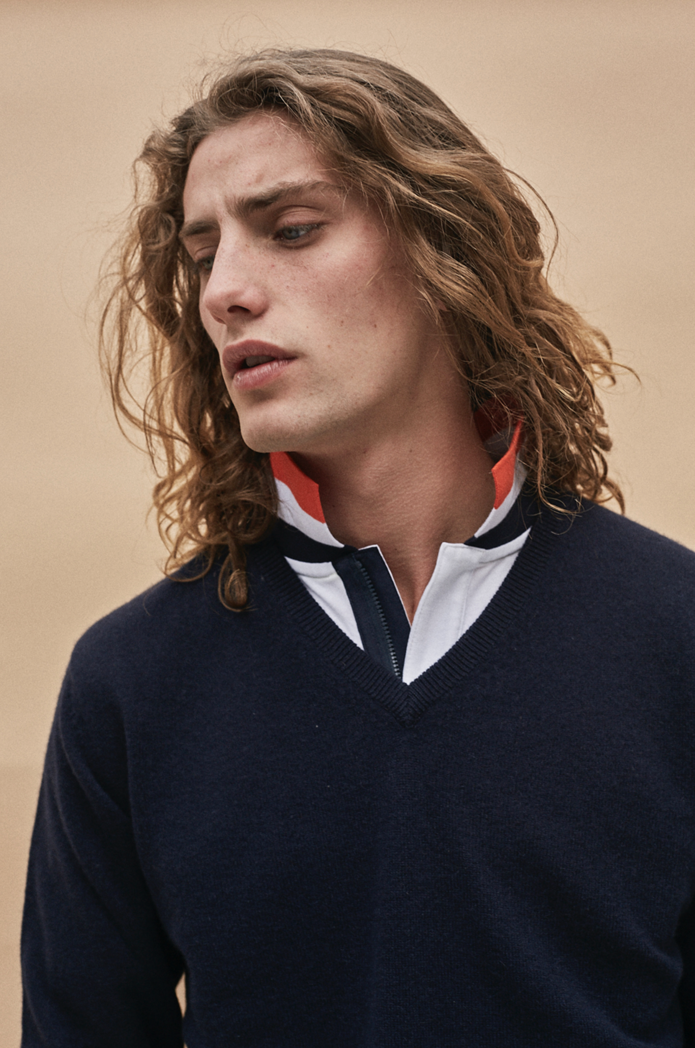 Joven con cabello rizado, rubio y largo, vestido con suéter azul oscuro con cuello y detalles en rojo y blanco, mirando hacia abajo y a la izquierda, fondo beige.