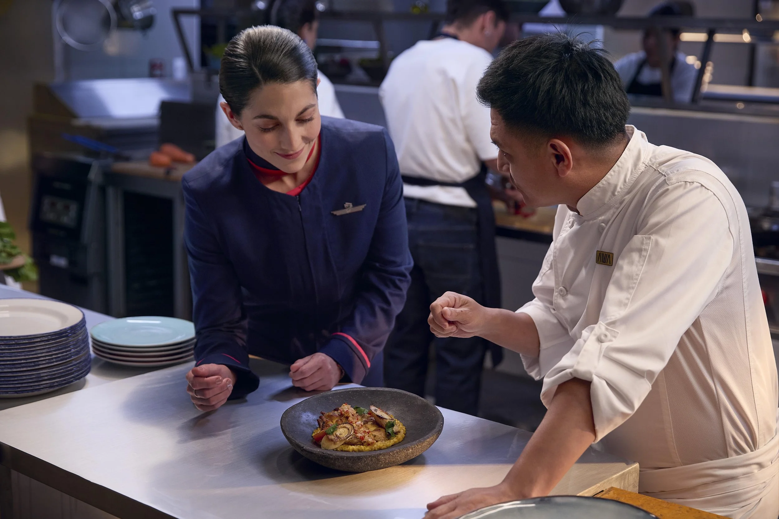 Un chef y una comensal observan un plato de comida en una cocina profesional.