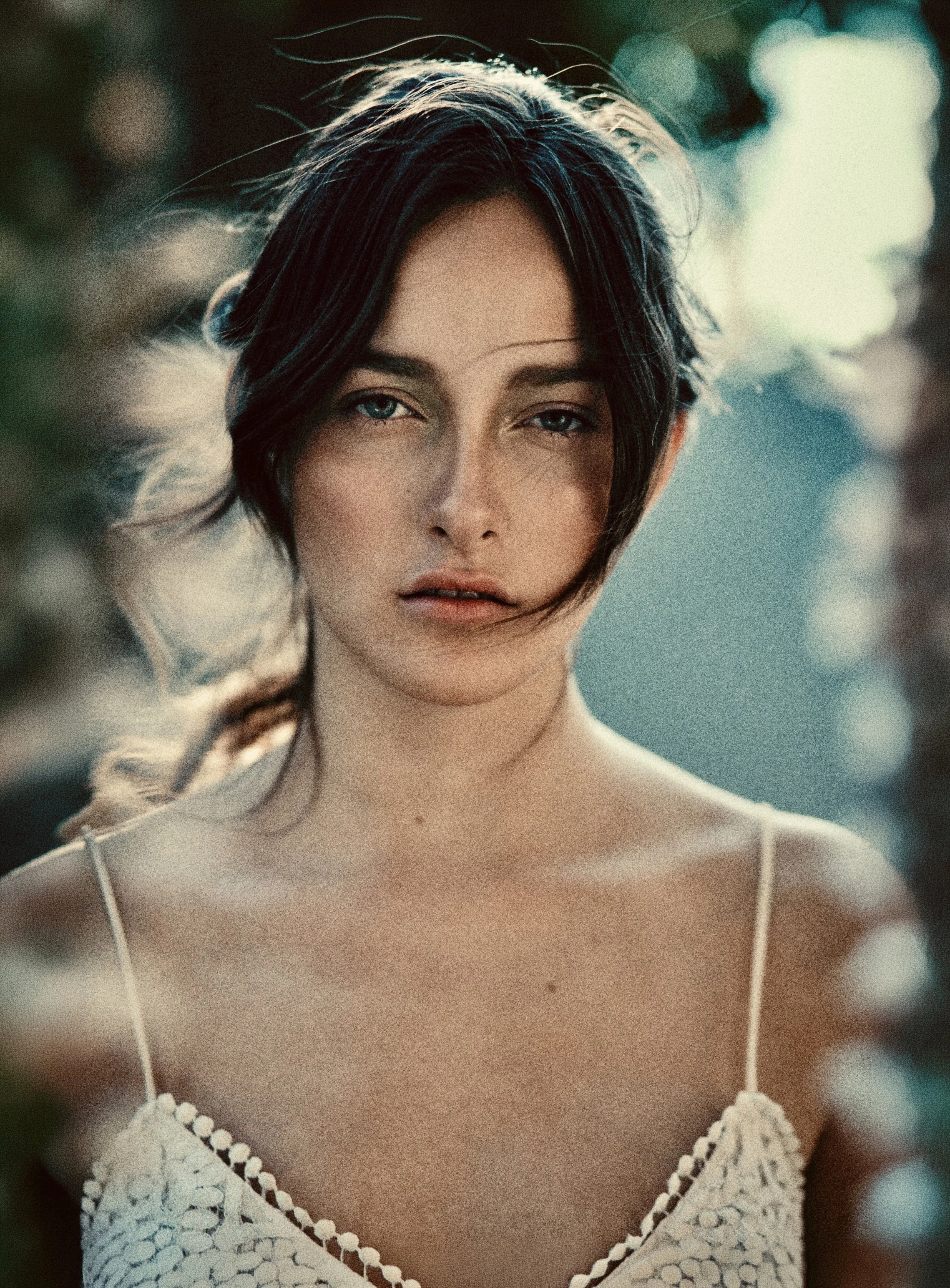 Retrato de una mujer joven con cabello oscuro y ojos azules, llevando un vestido blanco con detalles de encaje, en un entorno natural con fondo difuso y luz suave.