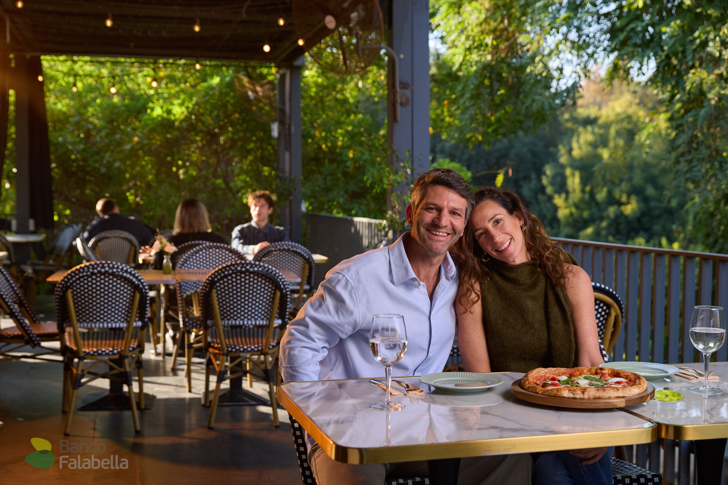 Pareja sonriendo en una terraza con cena, pizza y copas de agua, rodeada de árboles y luces decorativas.