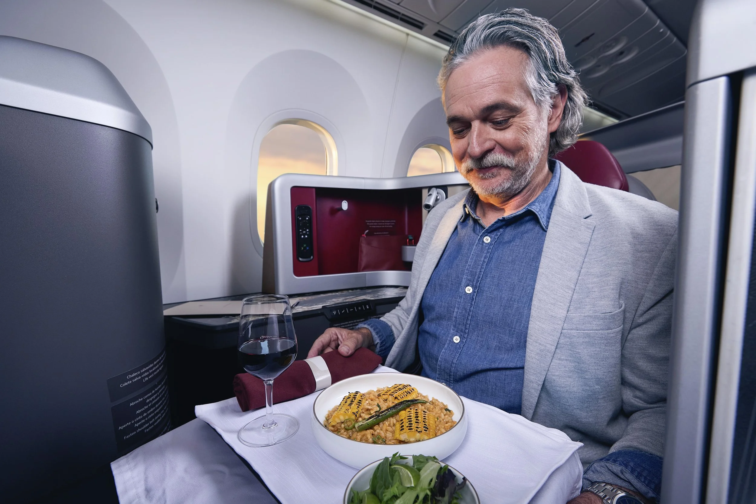 Hombre de cabello canoso y barba, vestido con blazer gris y camisa de mezclilla, sentado en un avión, disfrutando una comida con pasta que tiene calabaza en la parte superior, acompañado de una copa de vino tinto, con una ensalada de lechuga y lima e