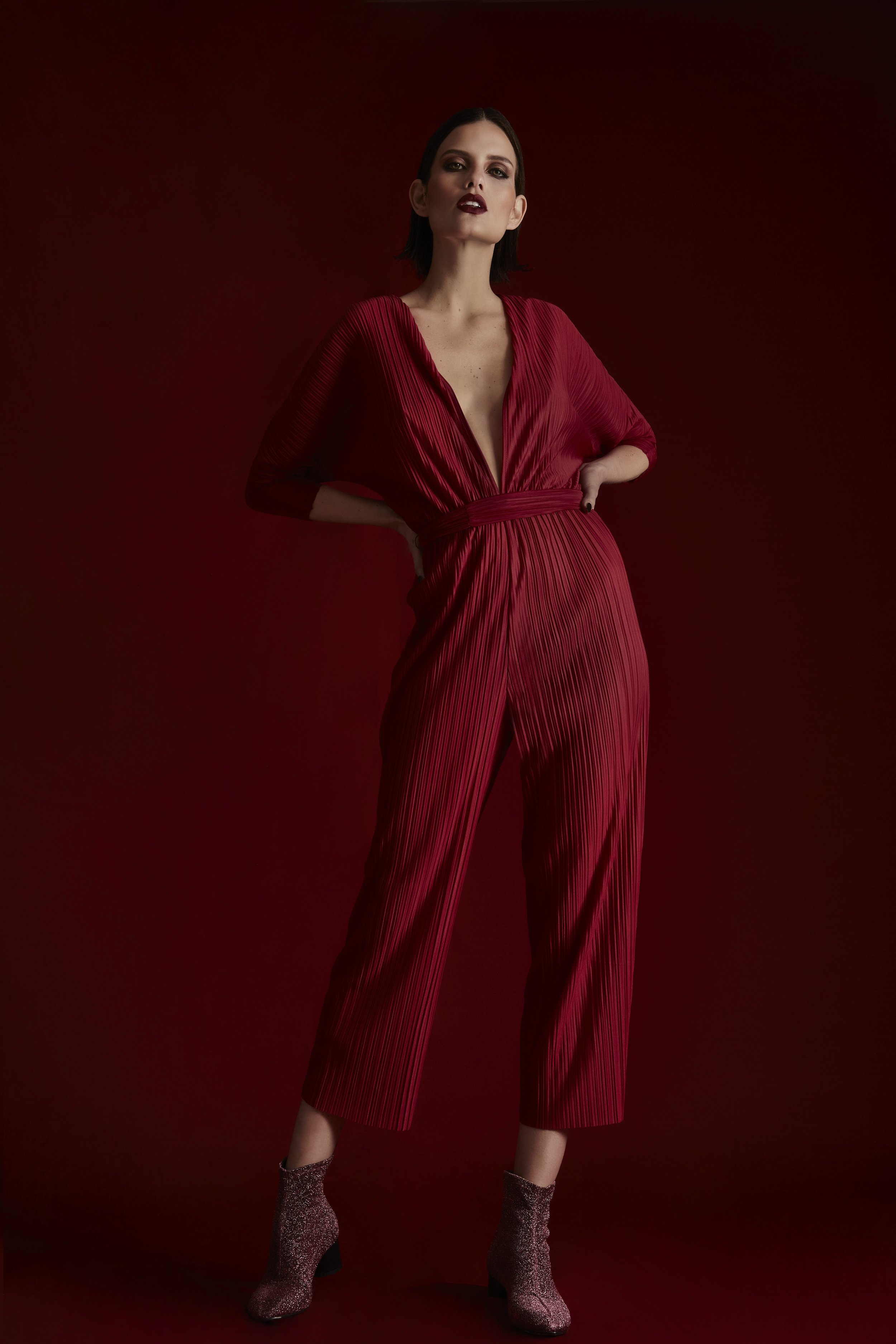 Mujer con vestido rojo brillante, de cabello oscuro y corto, posando con las manos en las caderas, fondo rojo oscuro.