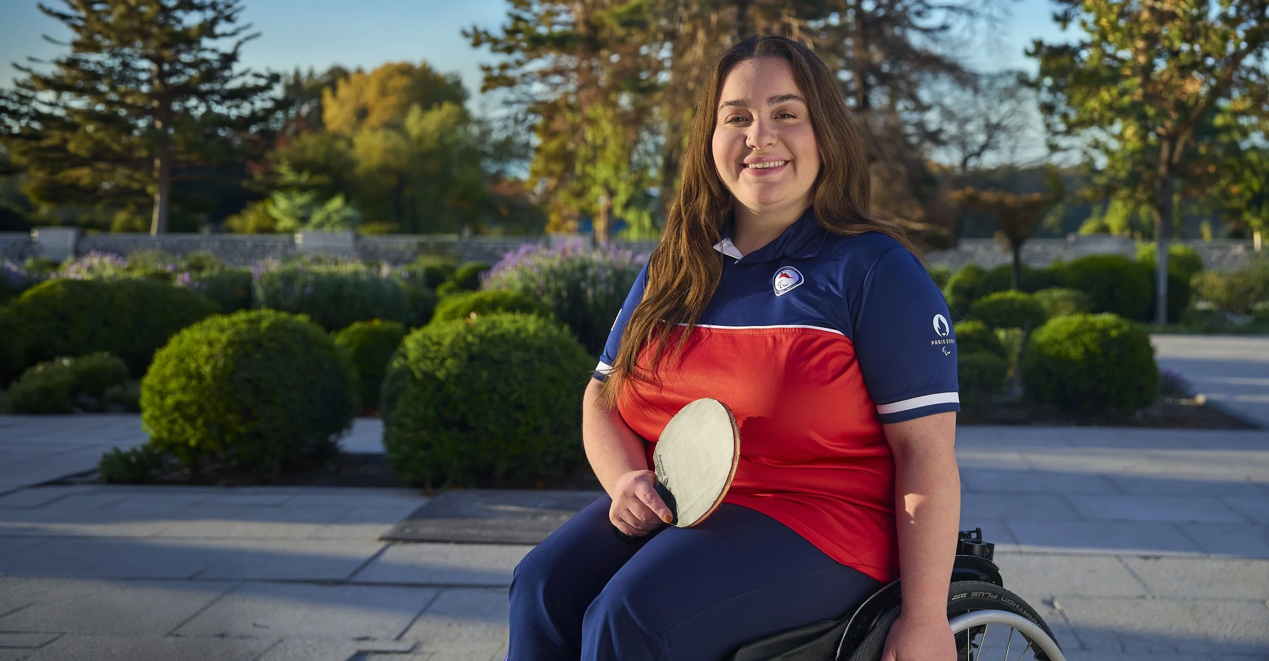 Juvenil con discapacidad en silla de ruedas, sonriente, con paleta de ping pong en mano, en un parque con árboles y arbustos verdes durante el día.