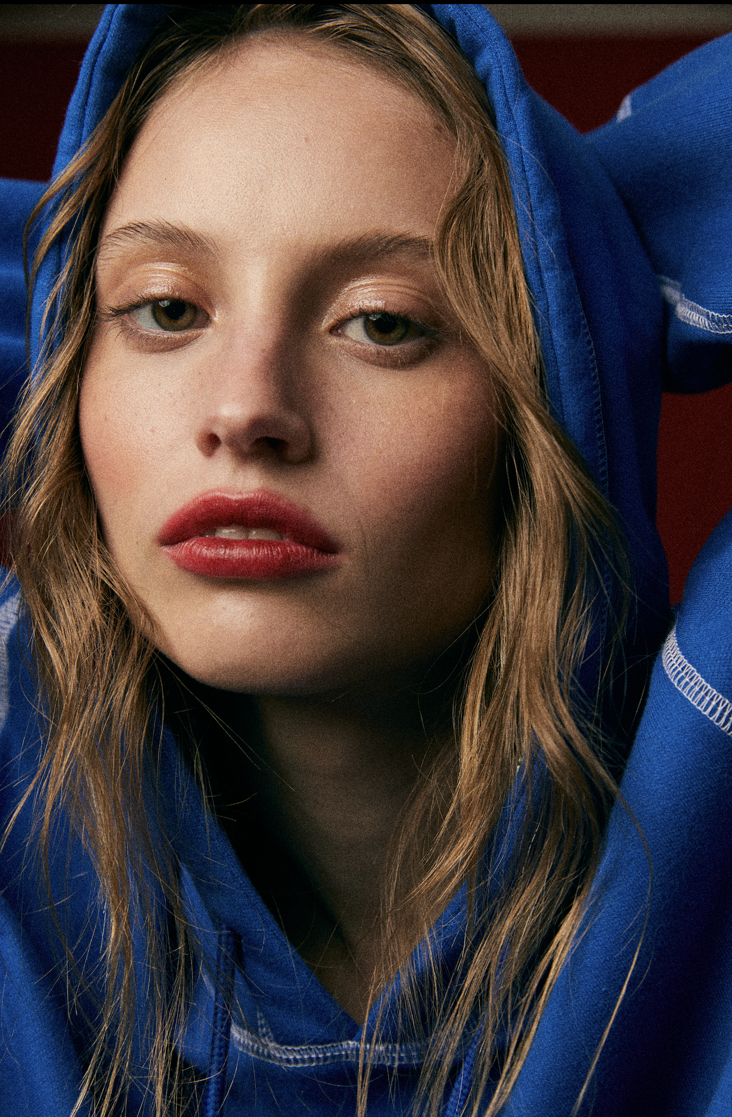 Retrato de una joven con cabello rubio, ojos verdes y labios rojos, usando sudadera azul y recargada con la cabeza hacia atrás.