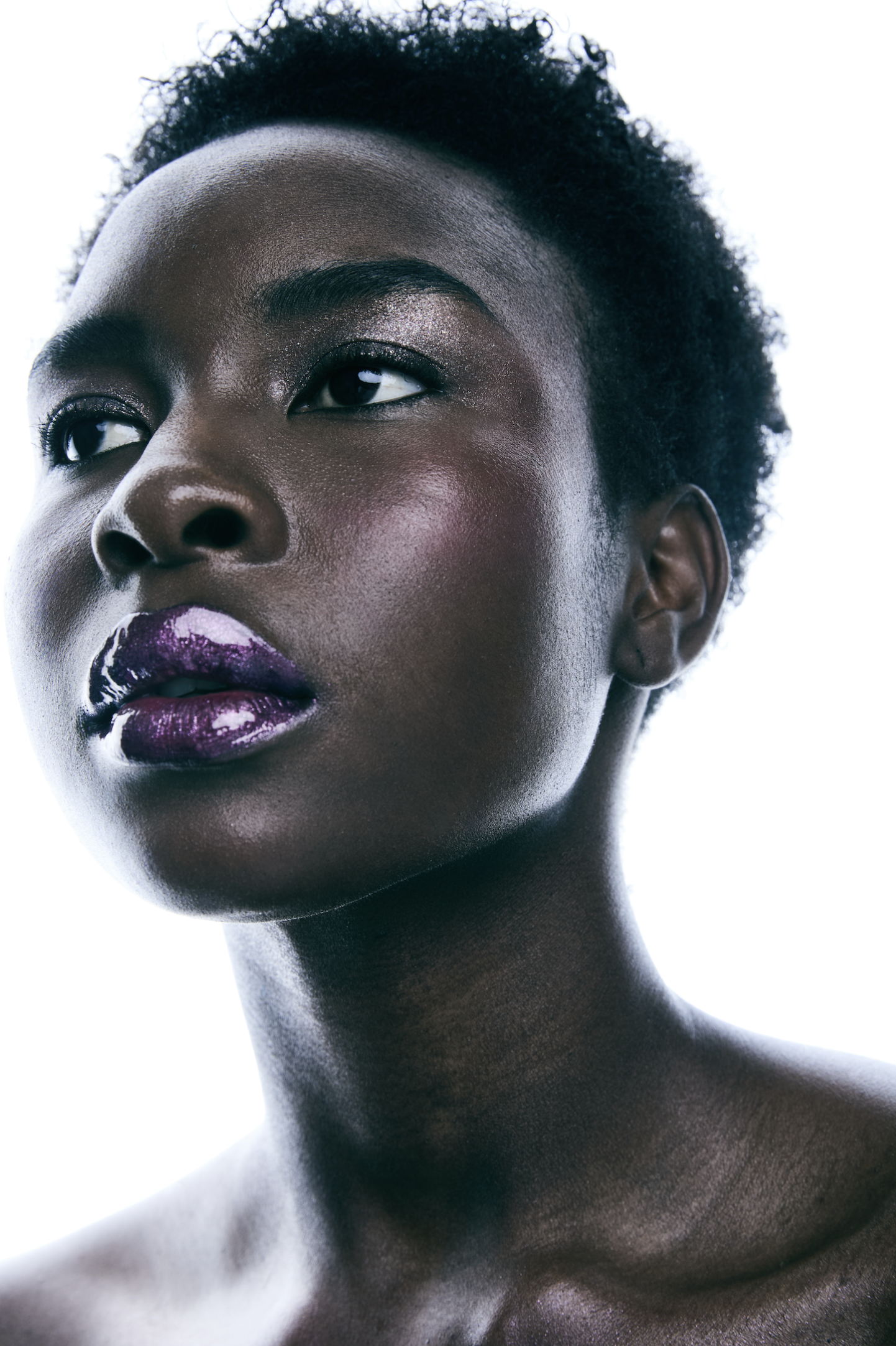 Primer plano de una mujer con piel oscura, ojos oscuros, peinado afro, labios morados y maquillaje en tonos metálicos en los ojos y mejillas, fondo blanco.