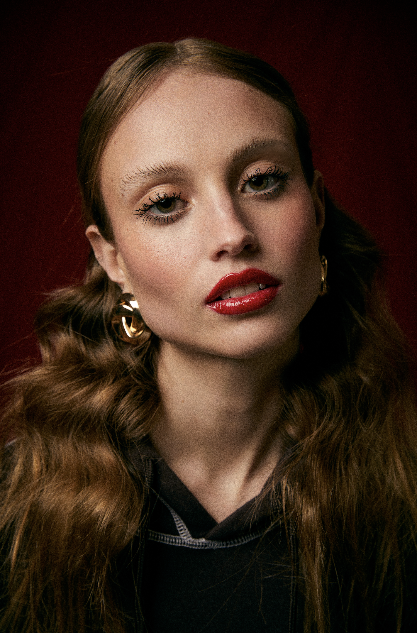 Retrato de una mujer con cabello rizado de color castaño claro, piel clara, maquillaje con labios rojos y ojos delineados, usando aretes dorados y ropa oscura, en fondo rojo oscuro.