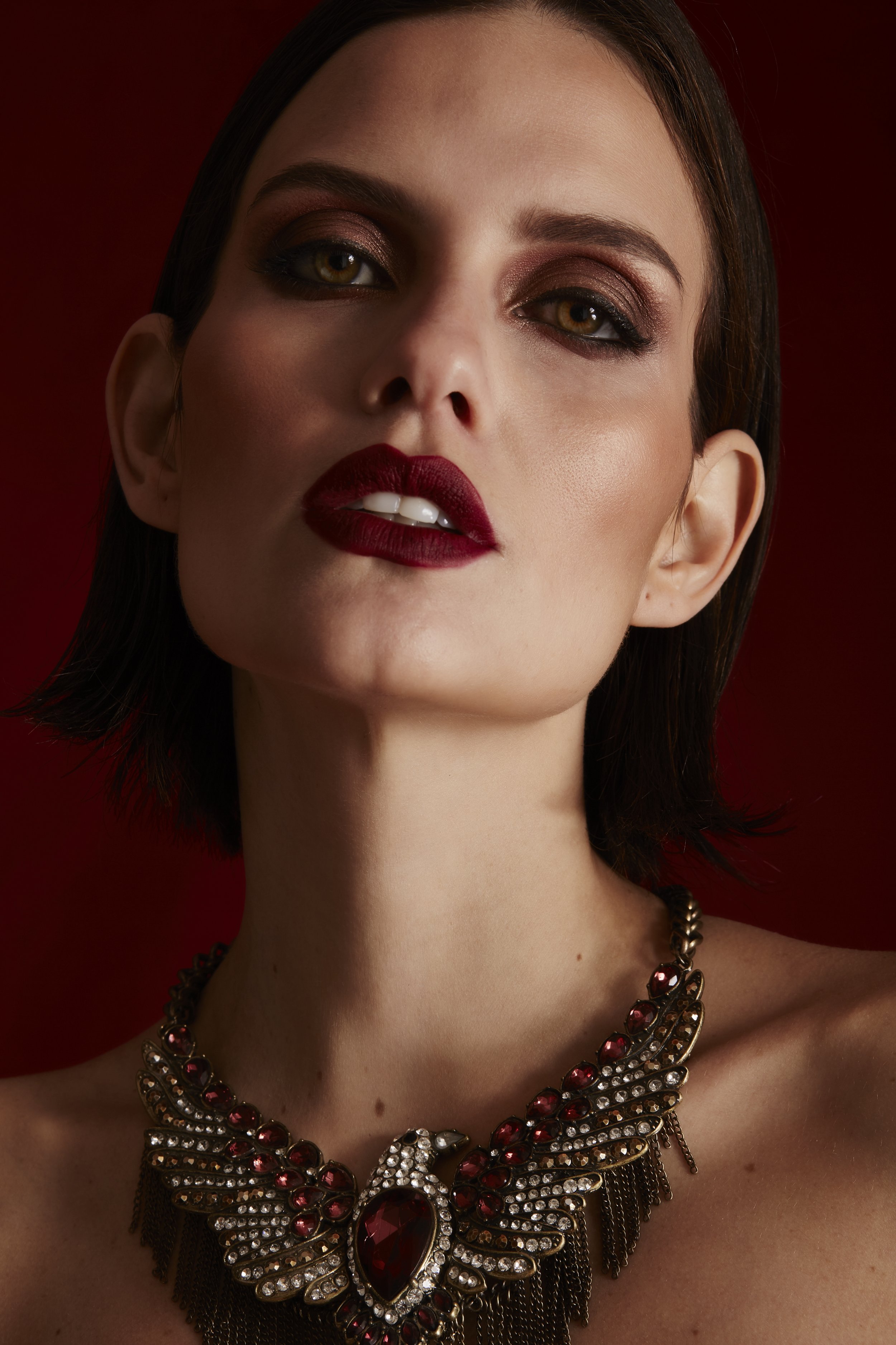 Primer plano de una mujer con maquillaje de ojos y labios oscuros, usando un collar grande con piedras rojas y detalles en plateado y negro.