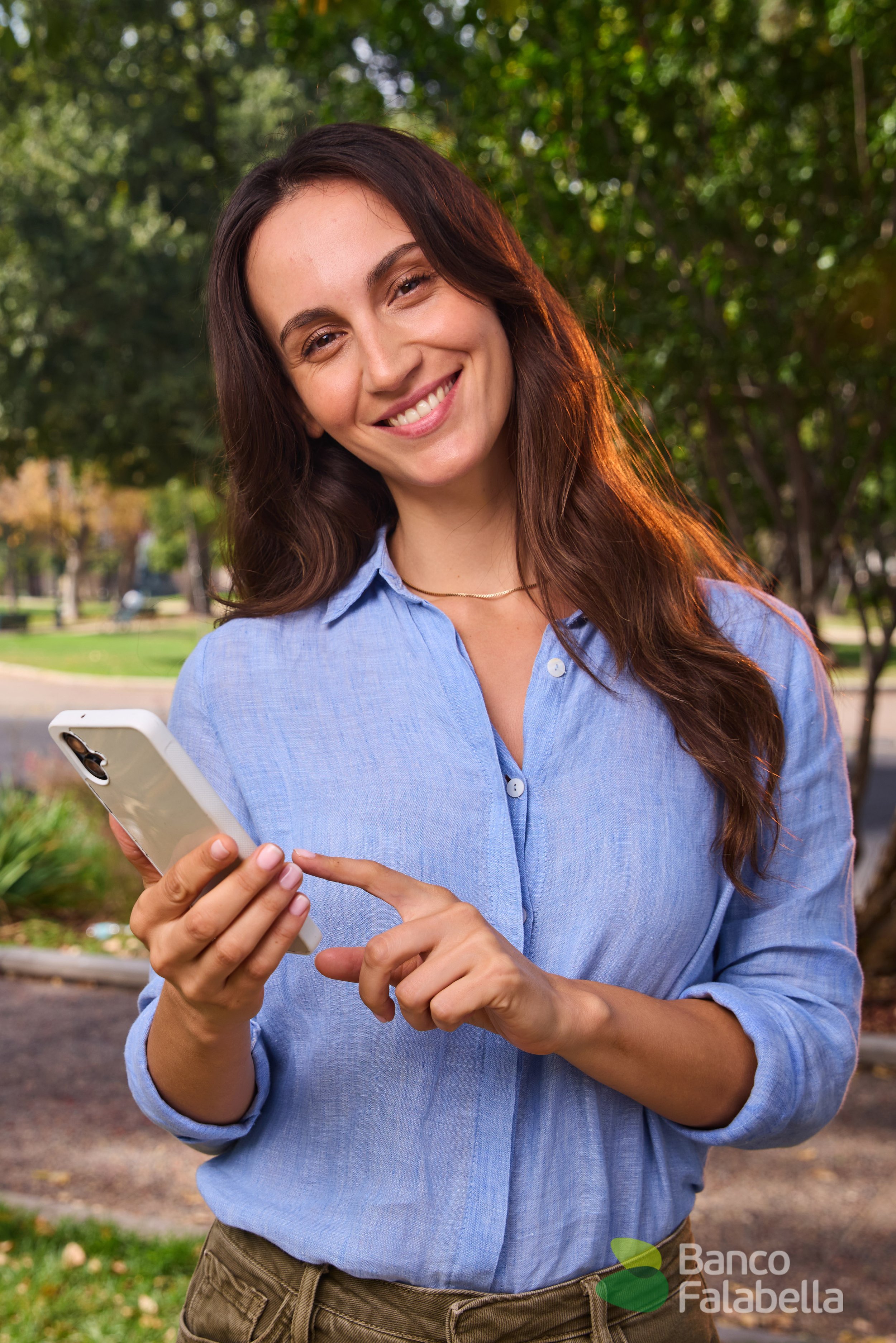 Joven mujer sonriente con camisa azul revisando su teléfono móvil en un parque con árboles verdes.