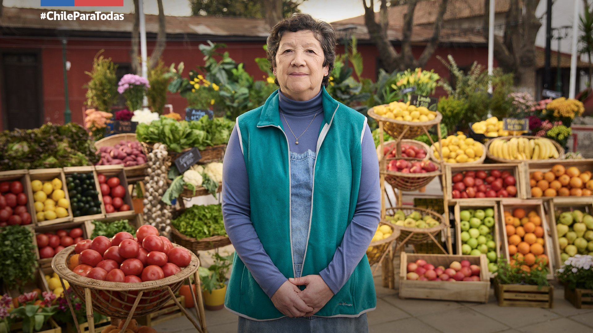 Mujer mayor en un mercado de frutas y verduras, con varias canastas de frutas como manzanas, plátanos y tomates, y flores de fondo.
