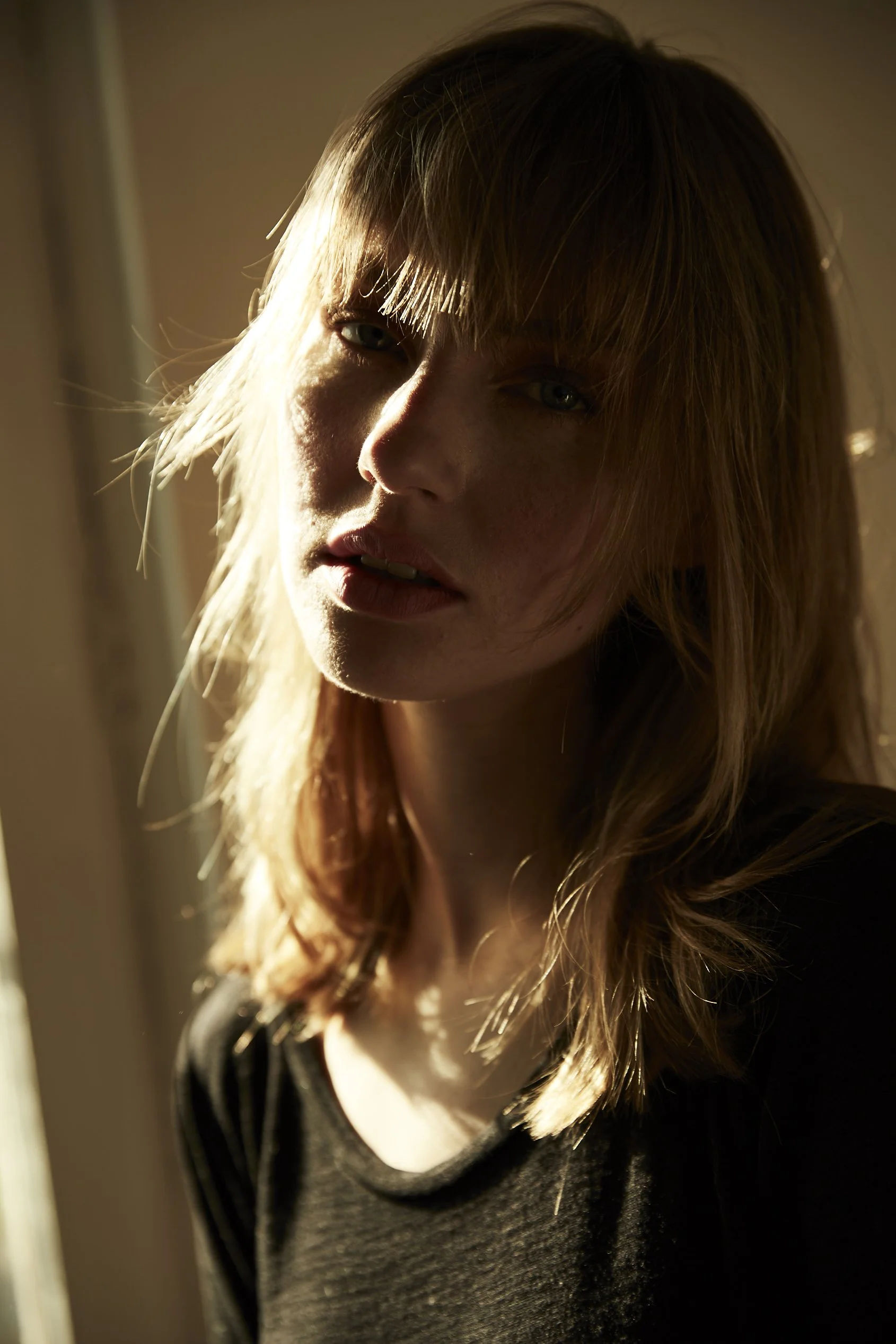 Retrato de una mujer con cabello rubio y largo, luciendo una expresión seria, con iluminación cálida y sombra en parte de su rostro, vestida con una camiseta oscura.